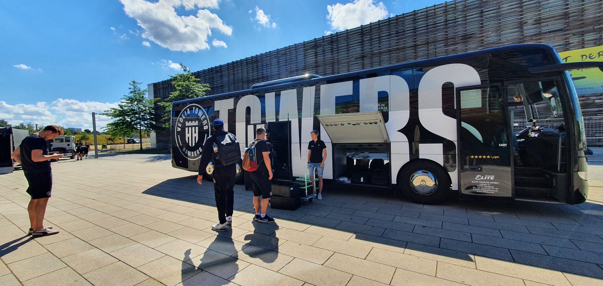 Menschen mit Gepäck vor einem schwarzen Reisebus mit großem Schriftzug 'TOWERS' und Logo 'VEOLIA TOWERS HAMBURG' bei sonnigem Wetter