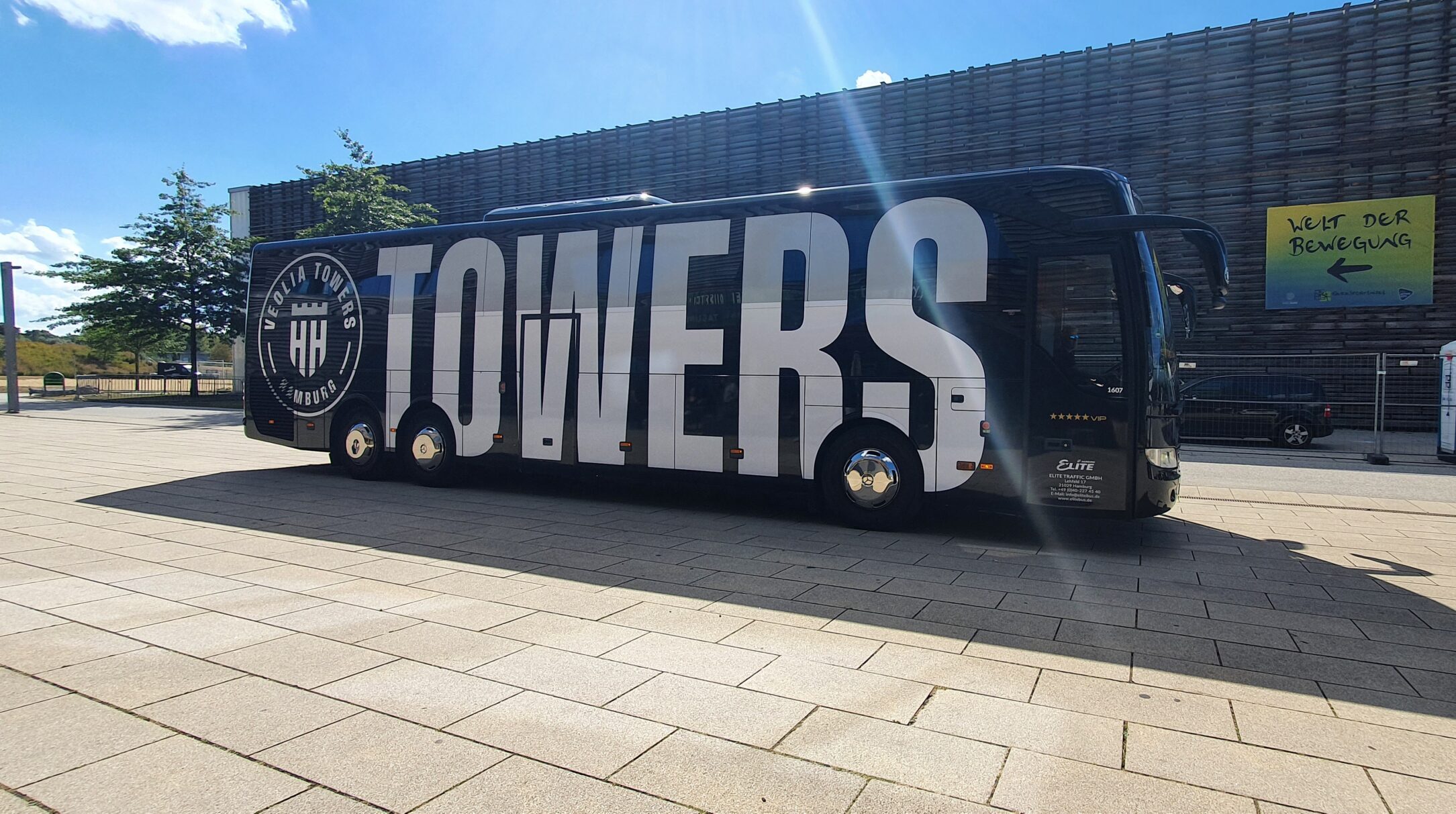 Schwarzer Reisebus mit großer weißer Aufschrift 'TOWERS' und rundem Logo 'Veolia Towers Hamburg' auf der Seite, geparkt auf gepflastertem Platz bei sonnigem Himmel