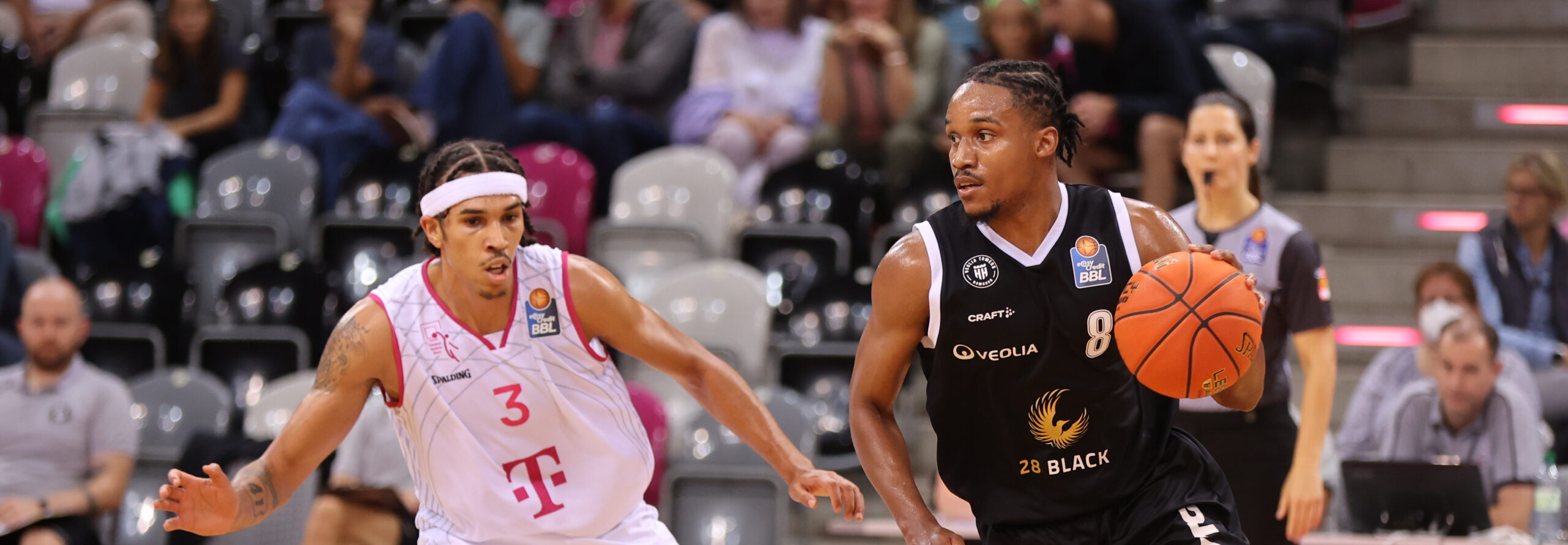 Zwei Basketballspieler in Aktion, einer in weißem Trikot mit der Nummer 3 und einer in schwarzem Trikot mit der Nummer 8, der den Ball dribbelt