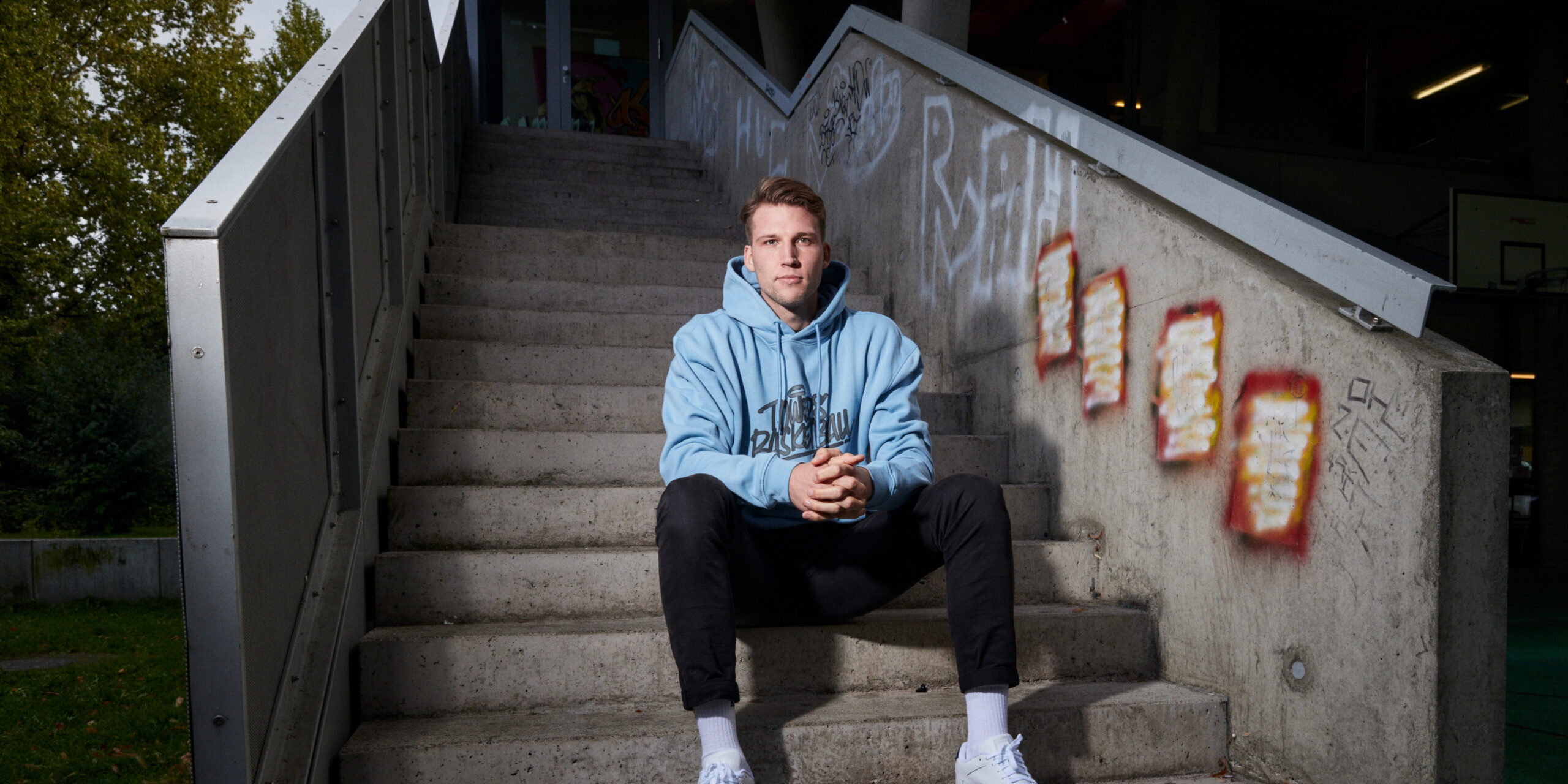 Junger Mann in blauem Kapuzenpullover sitzt auf einer Treppe mit Graffiti an der Wand, Hände vor dem Körper verschränkt
