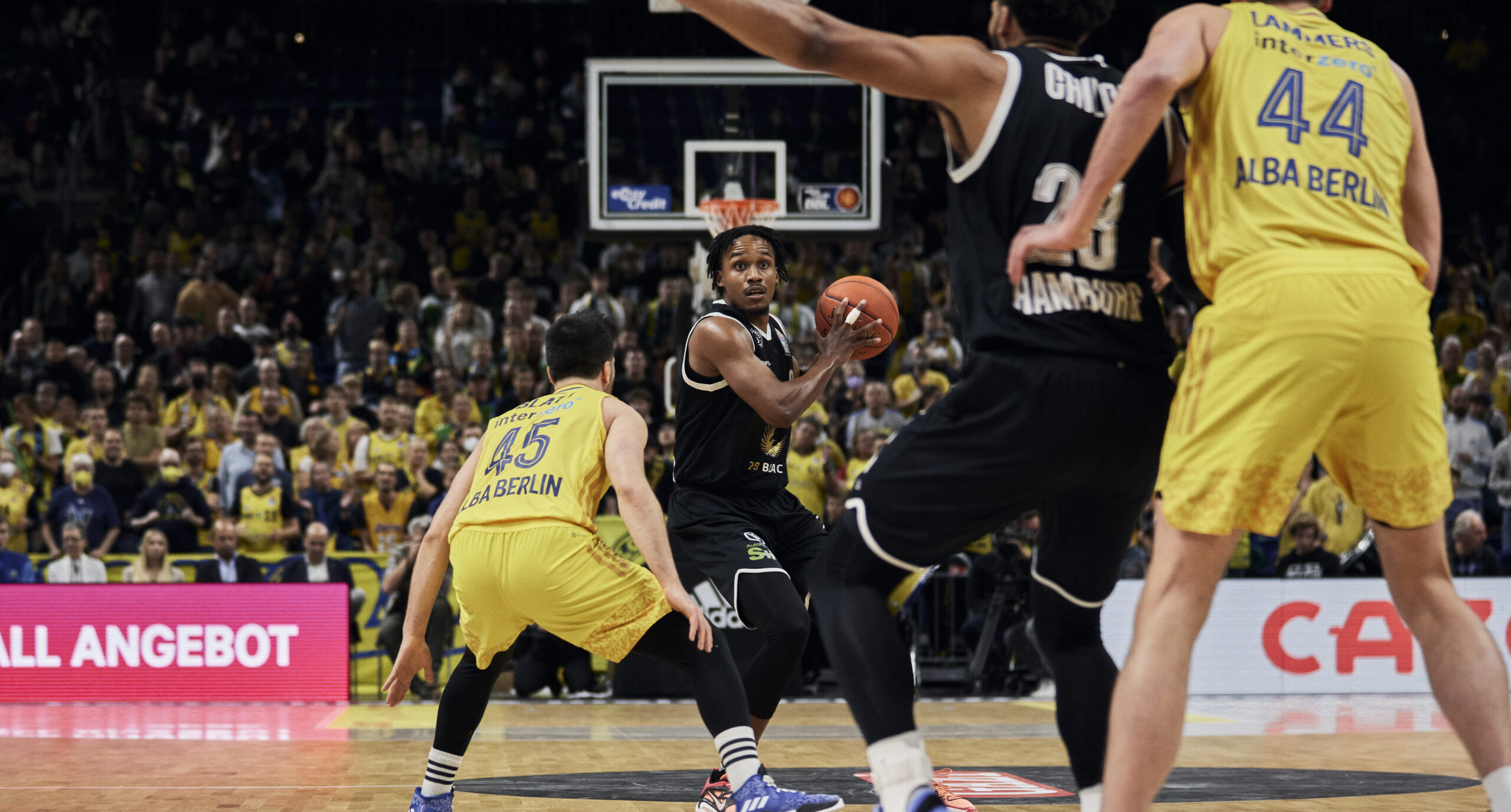 Basketballspieler in Aktion während eines Spiels zwischen ALBA Berlin und Hamburg Towers, ALBA-Spieler in gelber Uniform verteidigen gegen Spieler in schwarzer Uniform mit Ball vor dem Korb