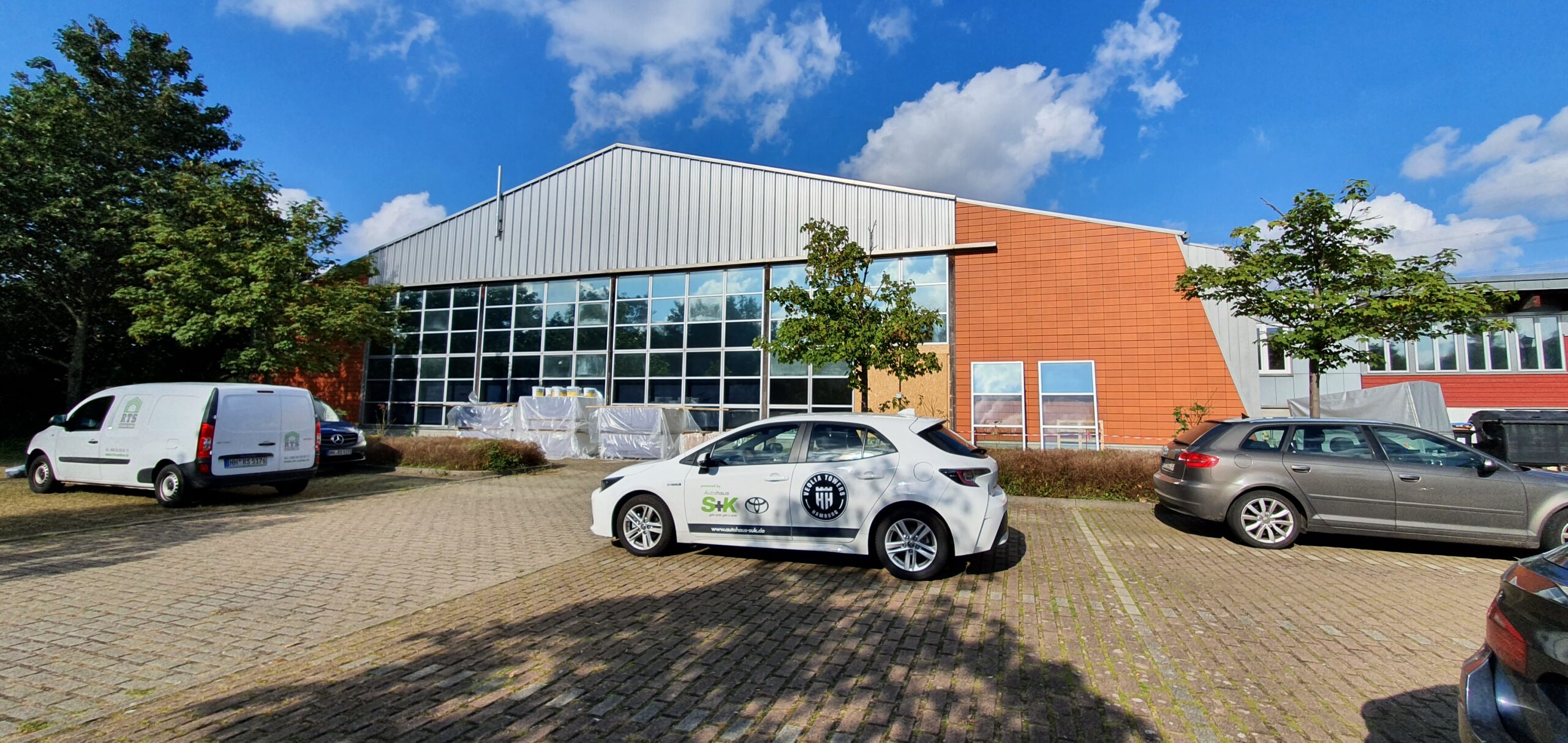 Parkplatz vor einem modernen Industriegebäude mit großen Glasfenstern und oranger Fassade, mehrere geparkte Autos, darunter ein weißer Toyota mit Firmenlogos