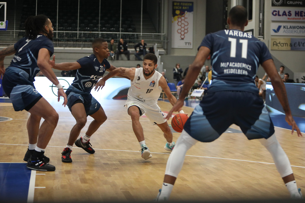 Basketballspieler in weiß dribbelt den Ball, umgeben von drei verteidigenden Spielern in blauen Trikots mit der Aufschrift 'Vargas' und 'MLP Academics Heidelberg'.
