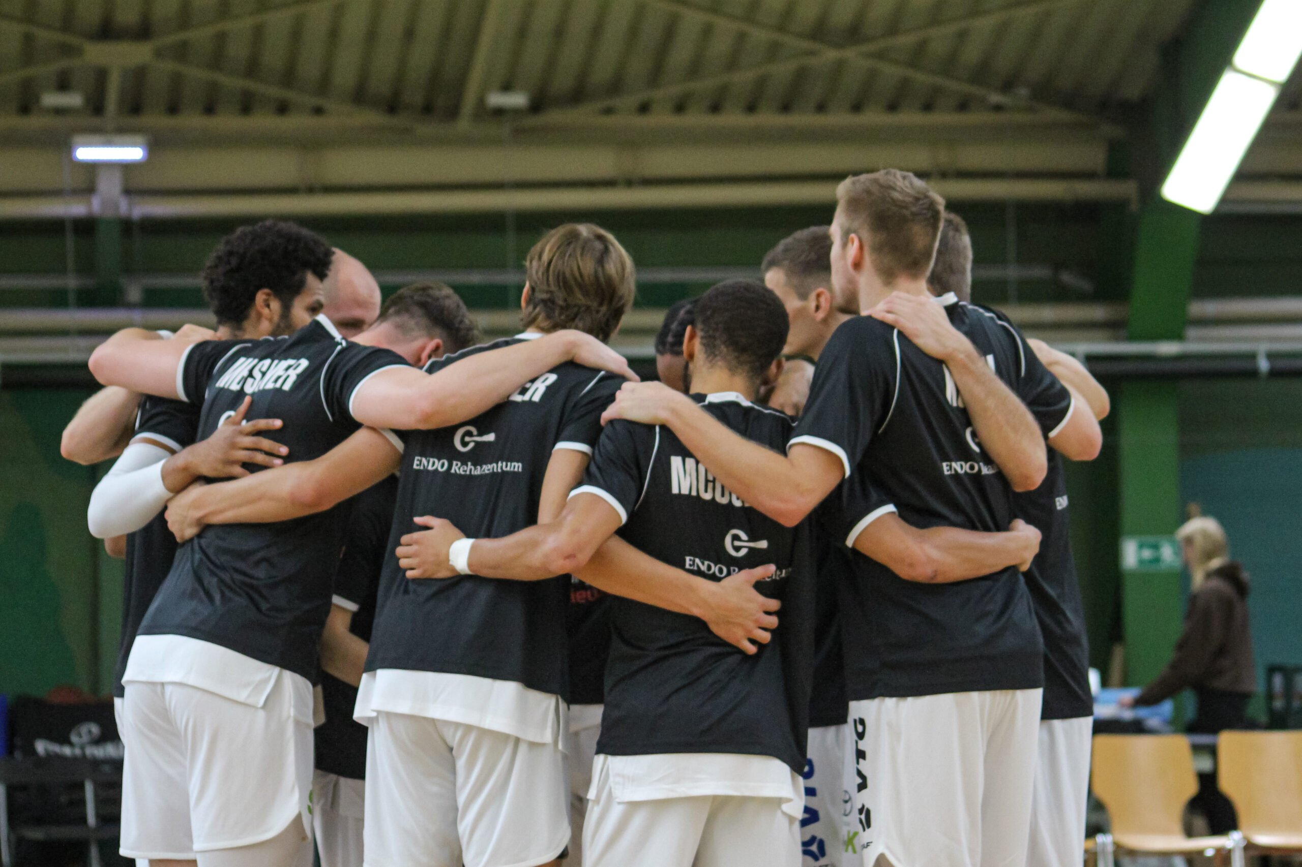 Basketballteam in schwarzen Trikots und weißen Hosen steht im Kreis und umarmt sich in einer Sporthalle