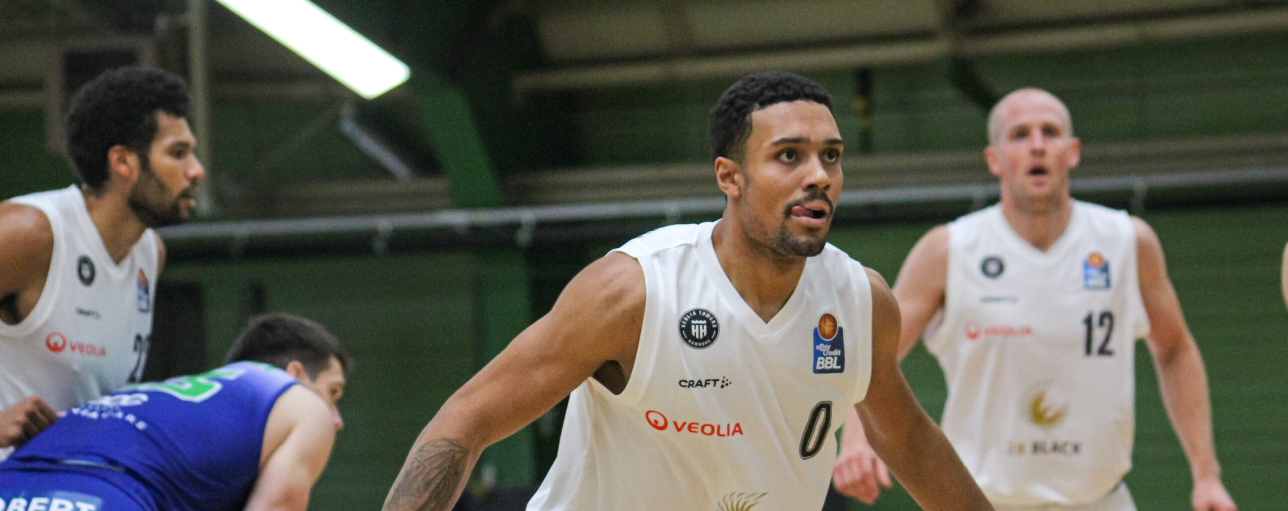 Drei Basketballspieler in weißen Trikots mit Nummern 0 und 12 in Aktion auf dem Spielfeld, ein Spieler in blau gebückt