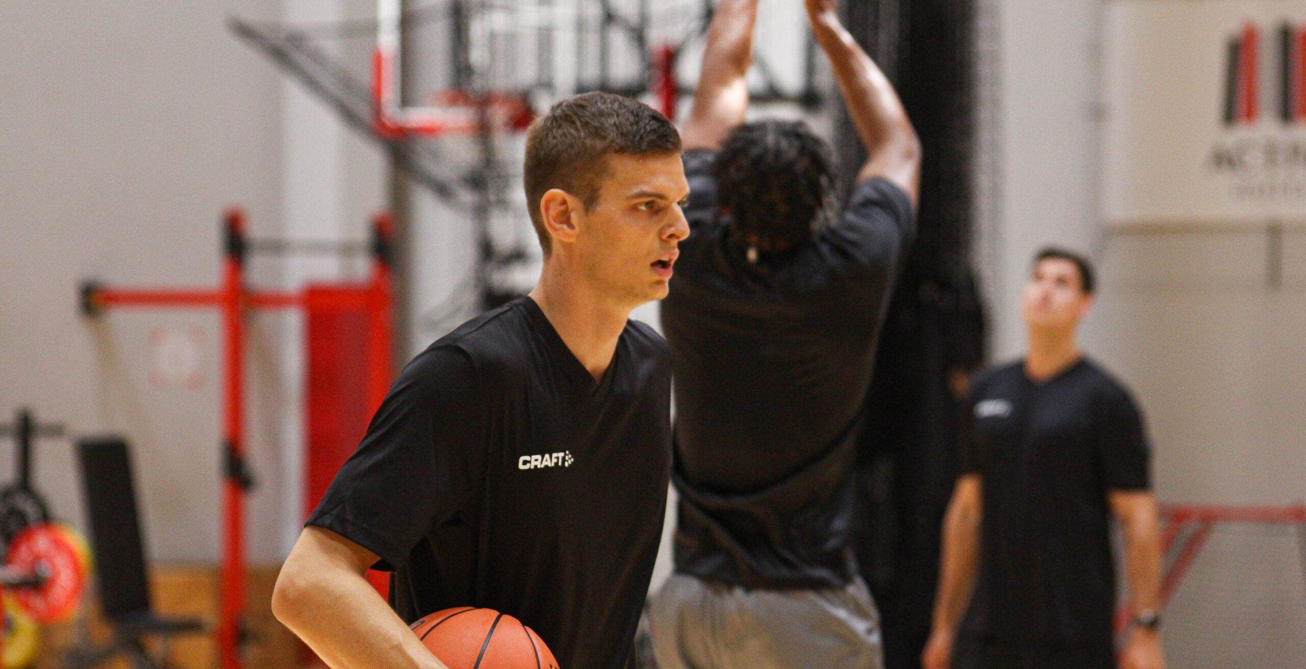 Junger Mann in schwarzem T-Shirt mit Basketball in der Hand in einer Sporthalle, im Hintergrund zwei weitere Männer beim Basketballspiel