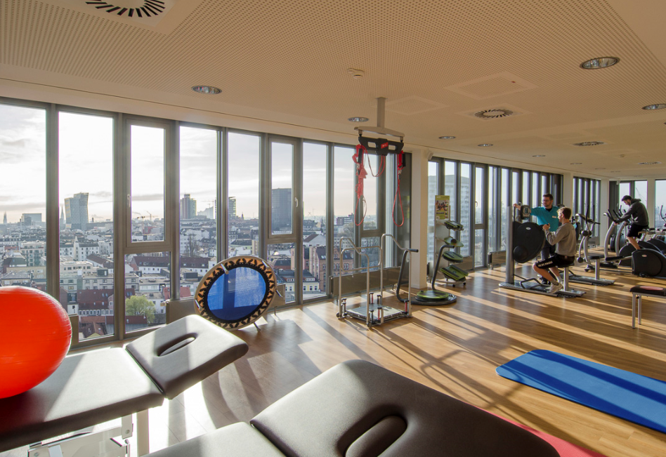 Fitnessraum mit großen Fenstern und Blick auf Stadt, Trainingsgeräte, Gymnastikmatte und zwei Personen beim Training