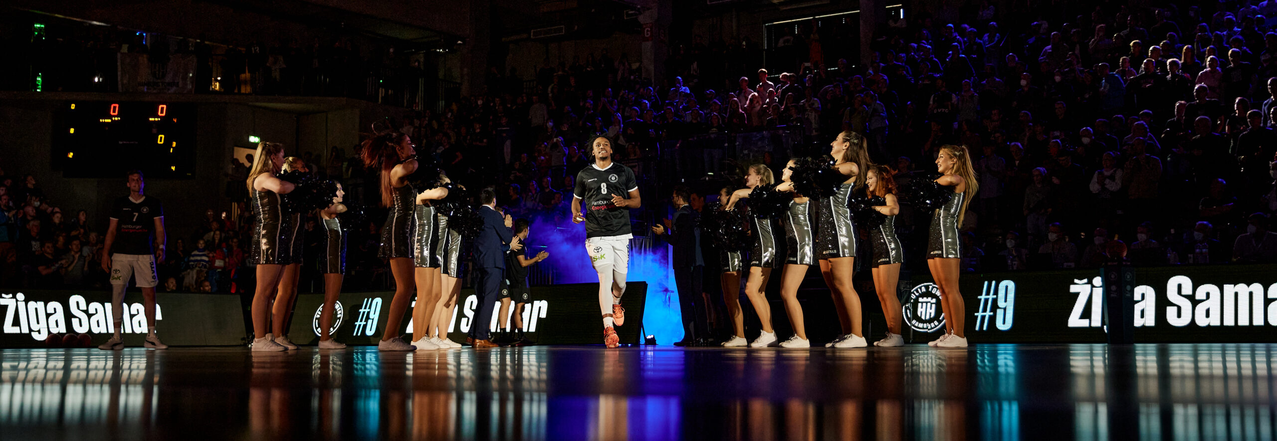 Basketballspieler läuft auf Spielfeld, flankiert von Cheerleadern in schwarzen Outfits, Zuschauer im Hintergrund