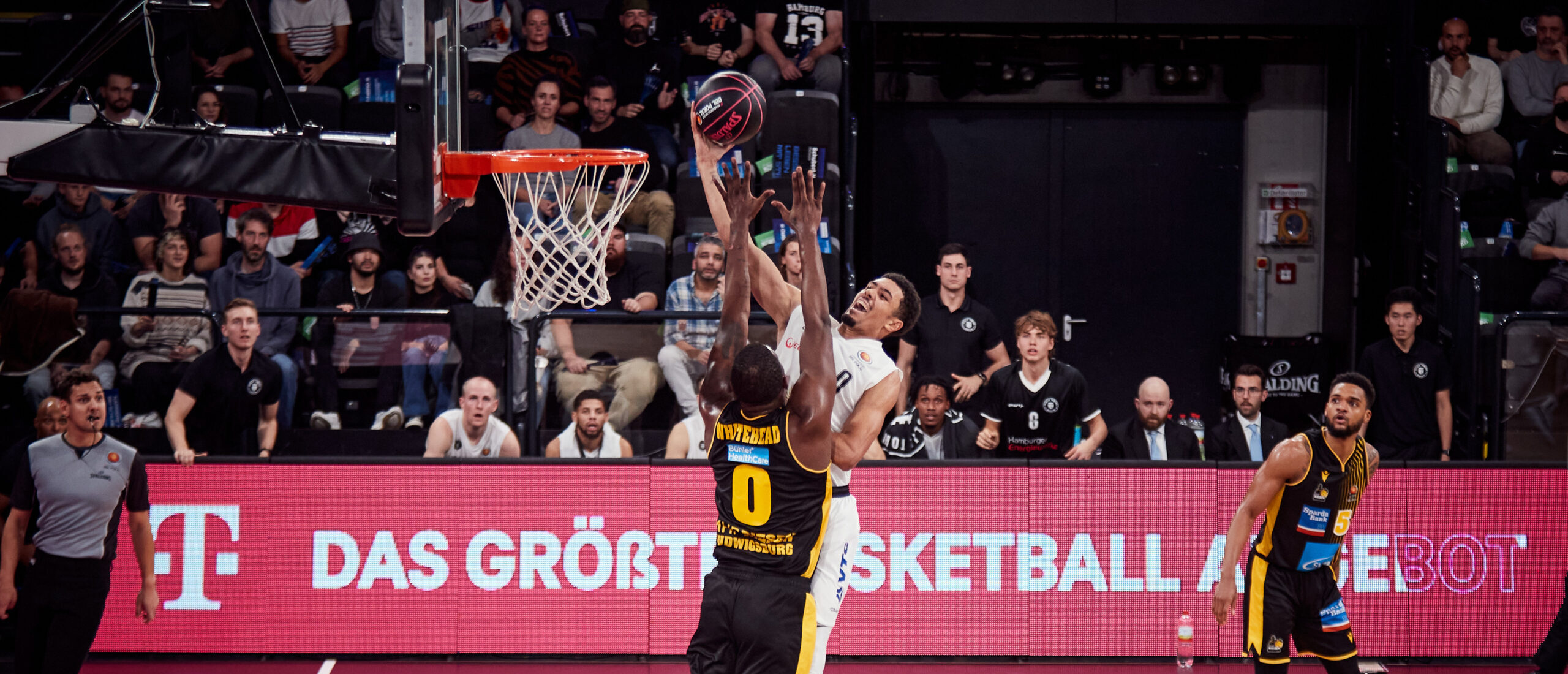Basketballspieler in weiß versucht Korb zu erzielen, verteidigt von Spieler in schwarzem Trikot mit gelben Akzenten, Zuschauer im Hintergrund