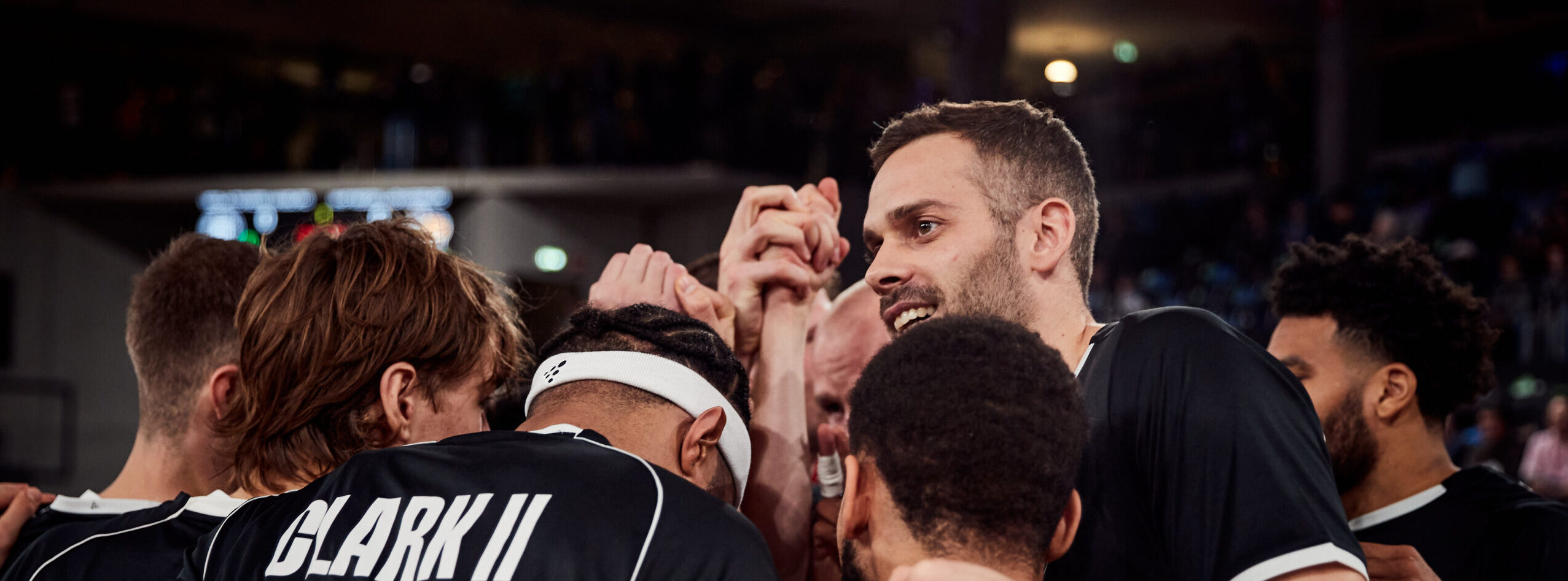 Basketballspieler in schwarzen Trikots bilden einen Teamkreis und legen die Hände zusammen