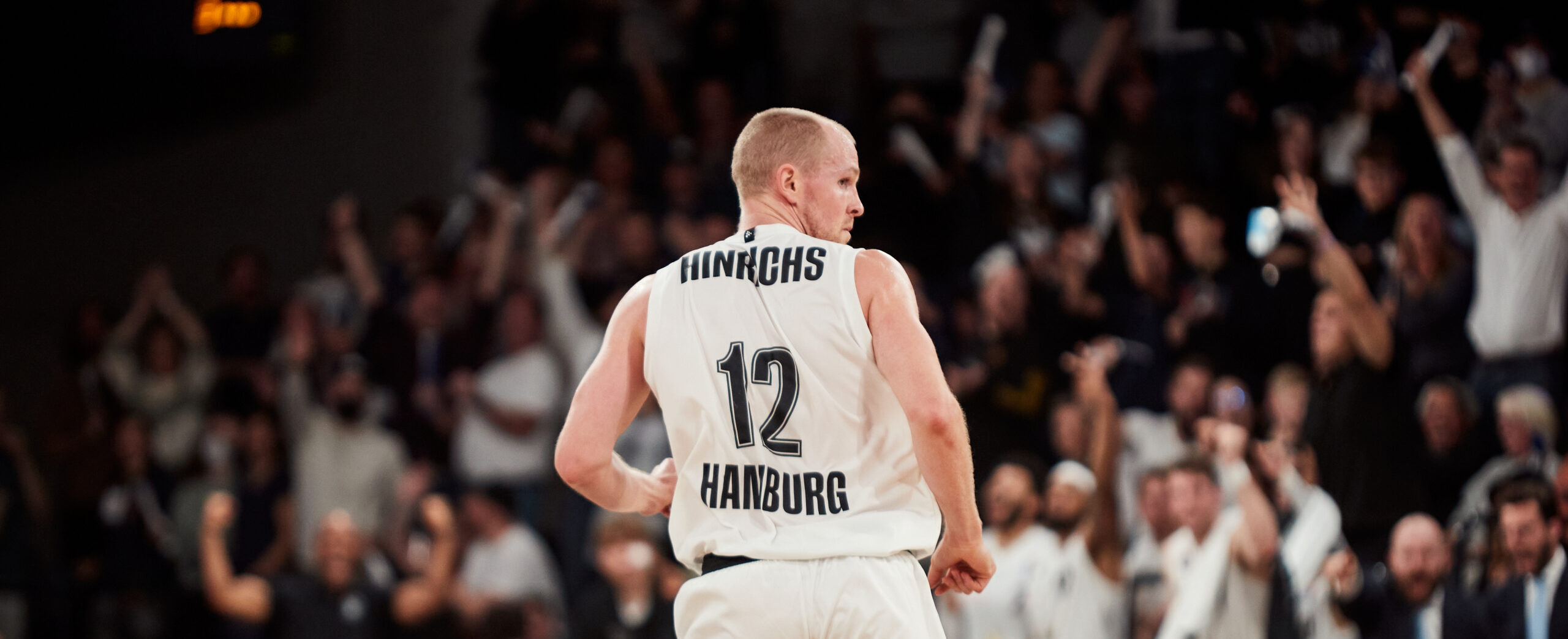 Basketballspieler mit Trikotnummer 12 und Aufschrift 'Hinrichs Hamburg' vor jubelndem Publikum