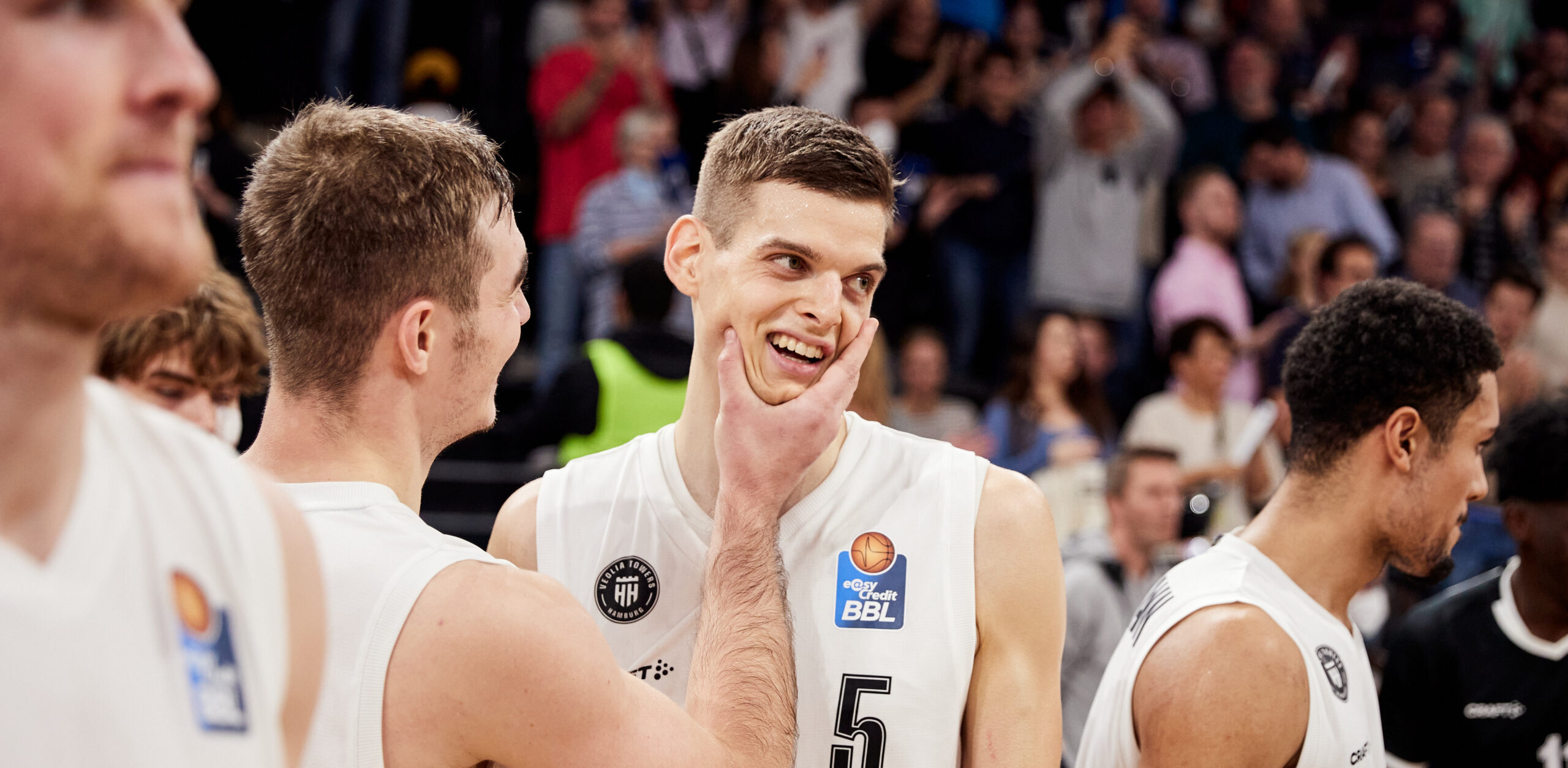 Basketballspieler in weißen Trikots mit der Nummer 5 lächelt, während ein Teamkollege ihm ins Gesicht greift, im Hintergrund Zuschauer
