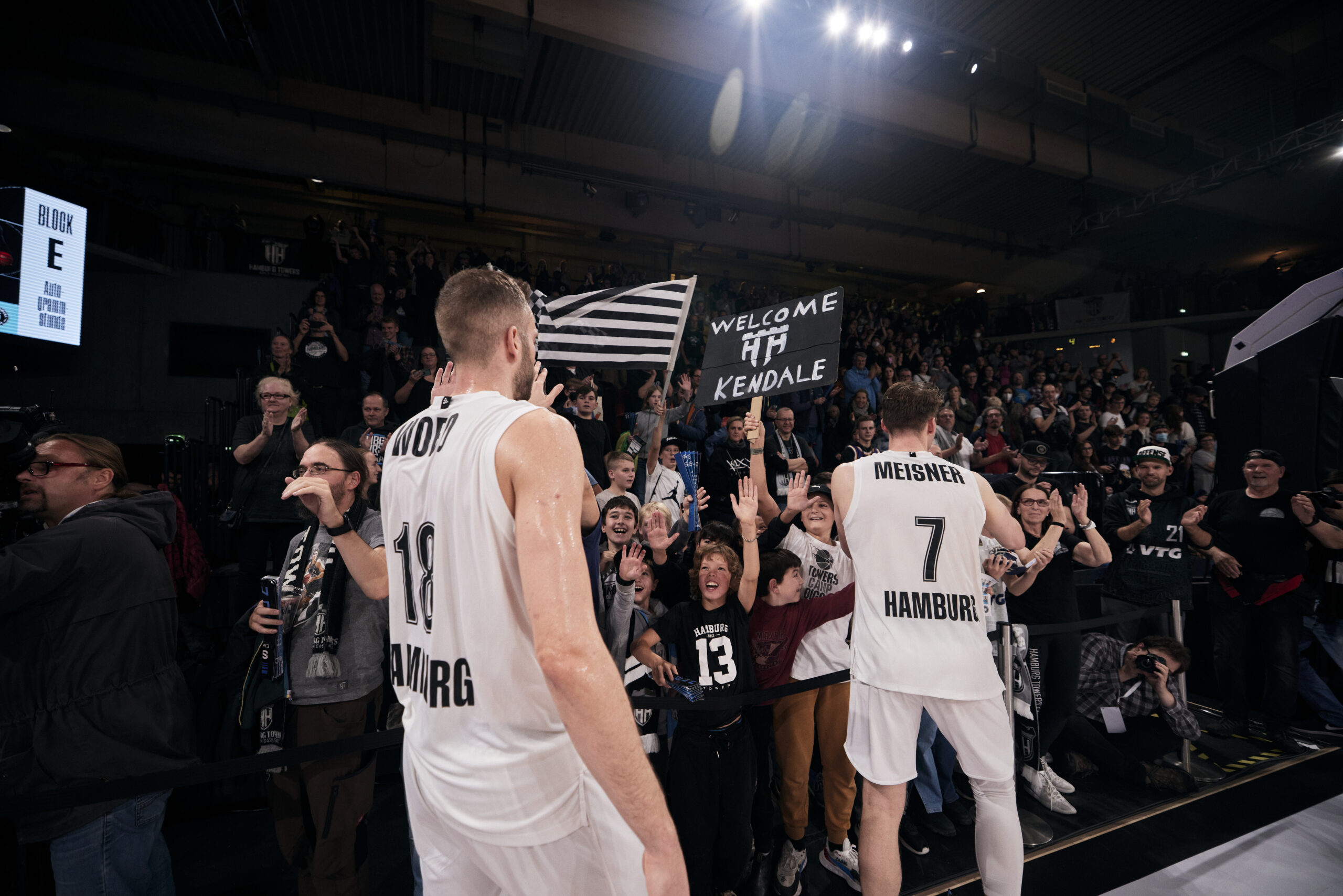 Zwei Basketballspieler der Hamburg Towers feiern mit jubelnden Fans in der Halle, eine Person hält ein Schild mit der Aufschrift 'Welcome Kendale'