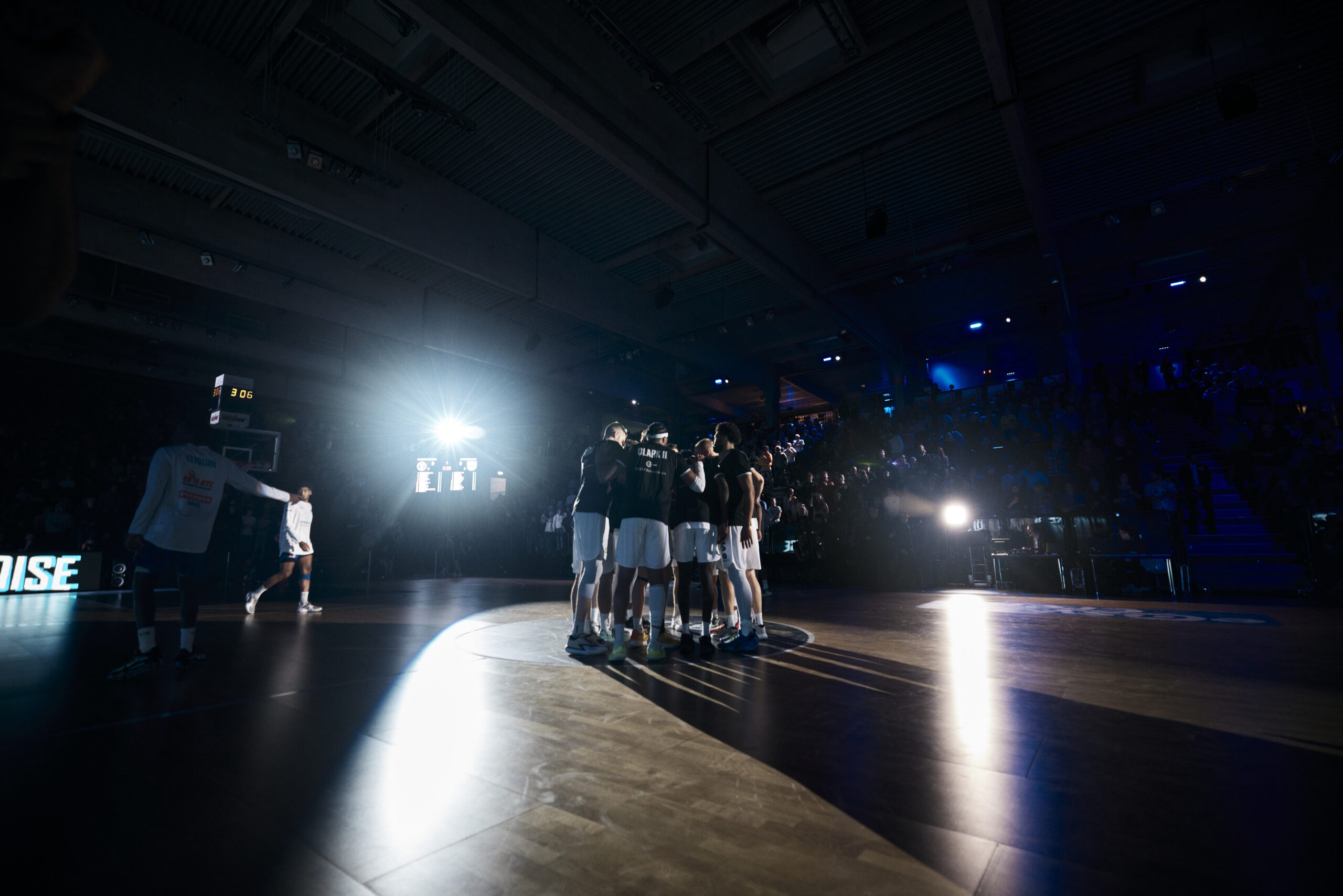 Basketballteam steht im Kreis auf dem Spielfeld, umgeben von dunkler Halle und starken Lichtquellen