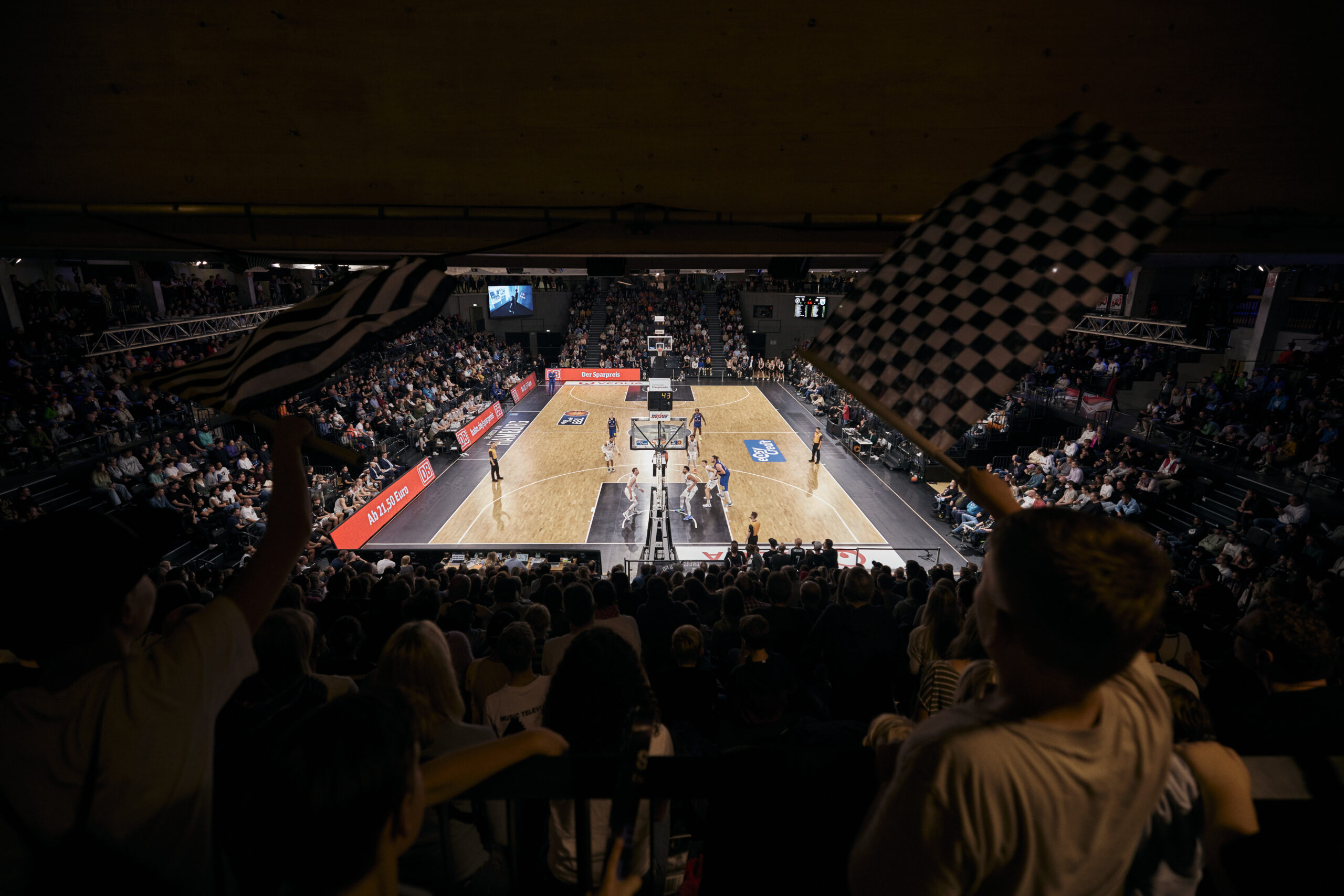 Basketballspiel in einer Halle mit Zuschauern, zwei Fans schwenken schwarz-weiße Fahnen im Vordergrund