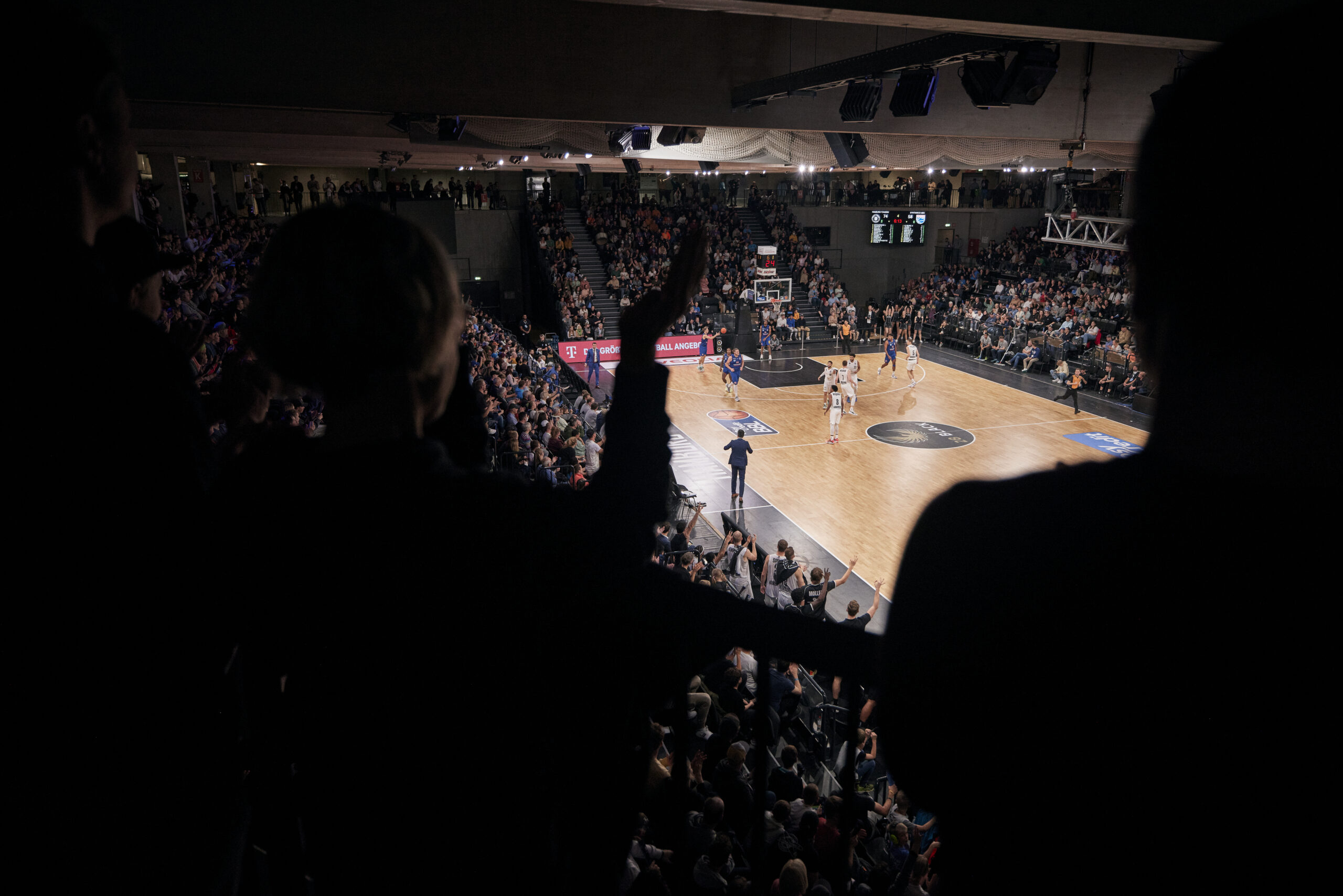 Basketballspiel in einer Halle mit Zuschauern, die von oben auf das Spielfeld blicken und jubeln