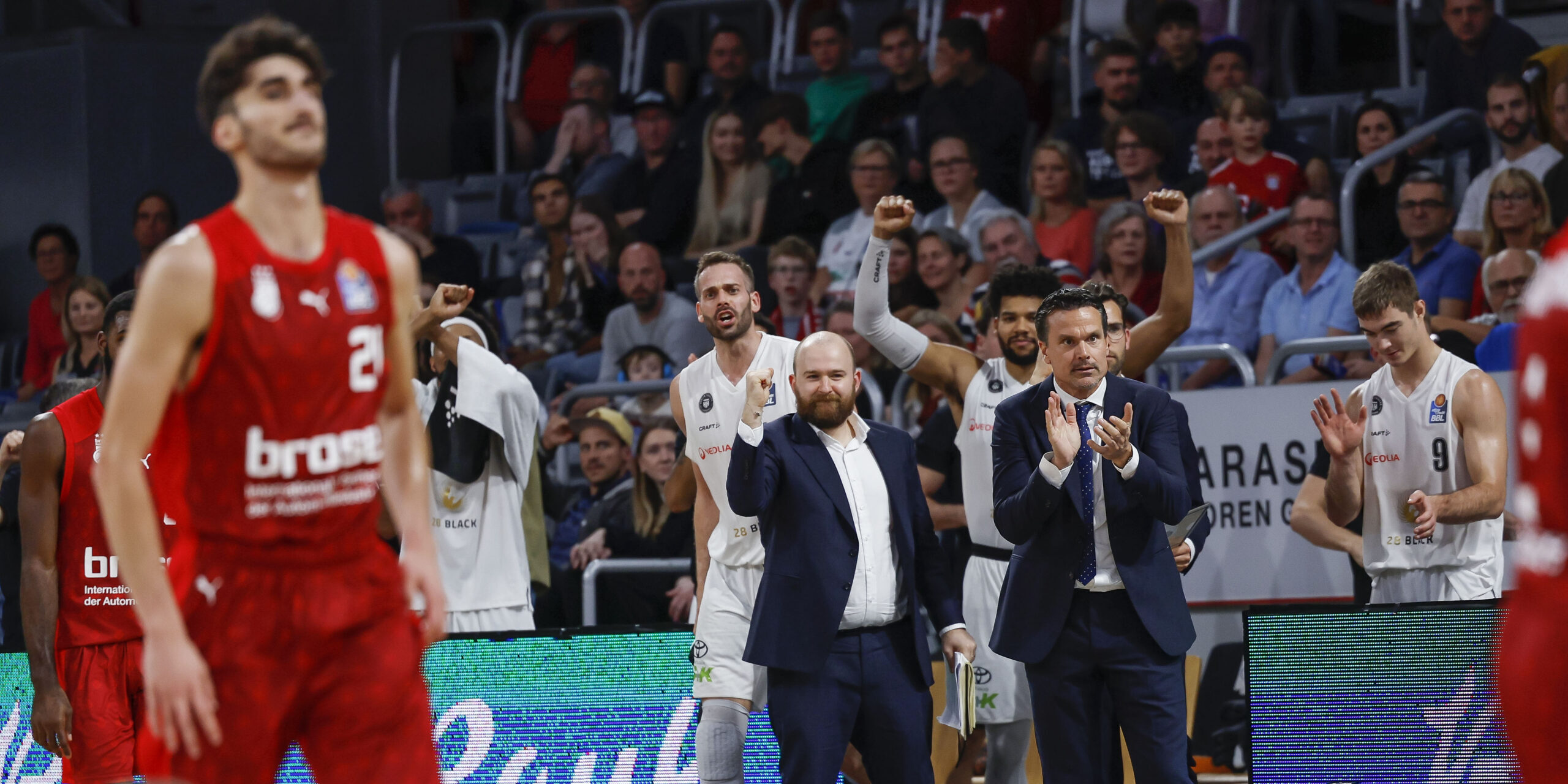 Basketballspieler in roten und weißen Trikots auf dem Spielfeld, Trainer in Anzügen klatschen, Zuschauer im Hintergrund