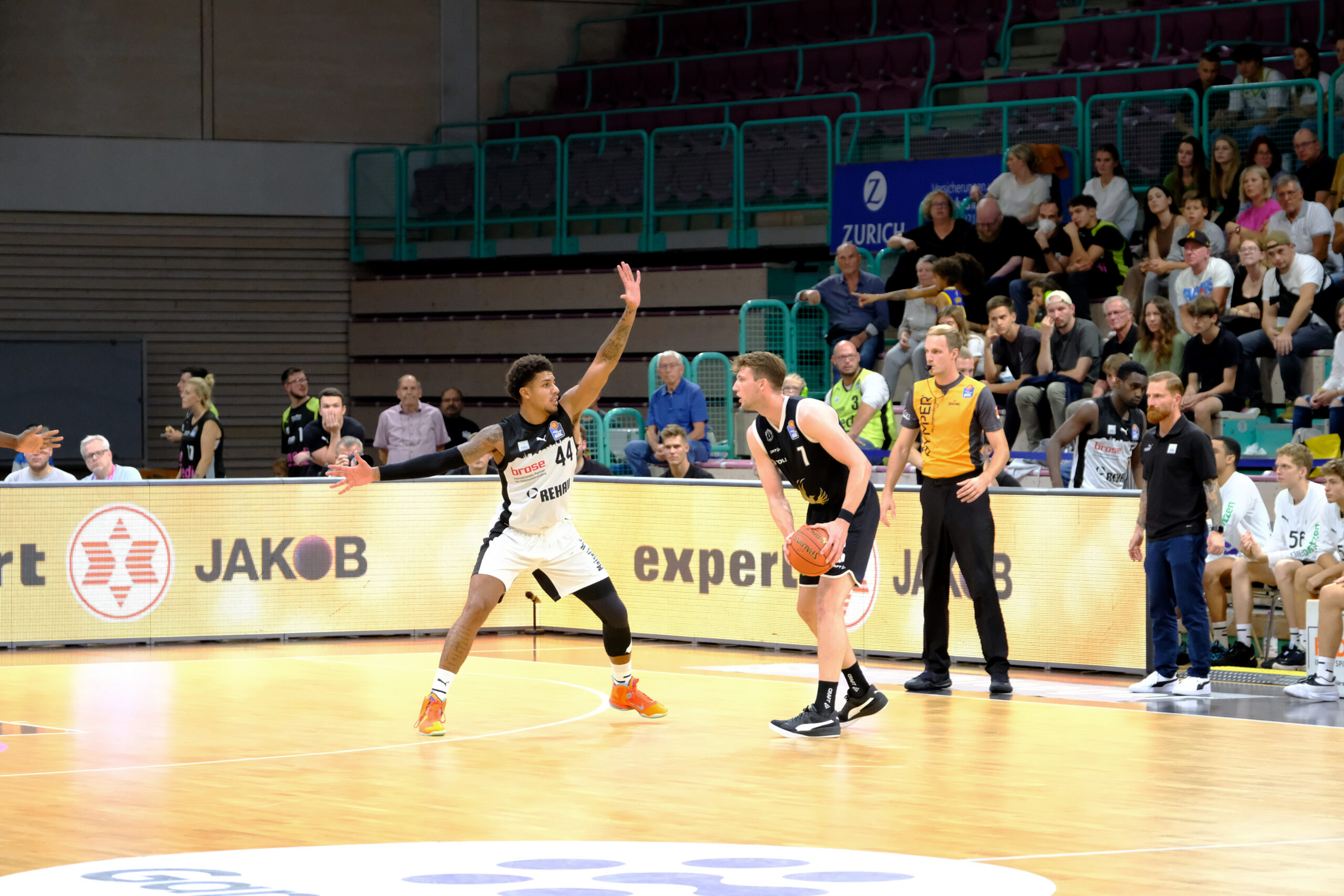 Basketballspieler in Aktion, einer verteidigt mit erhobenen Armen, der andere hält den Ball, Schiedsrichter und Zuschauer im Hintergrund