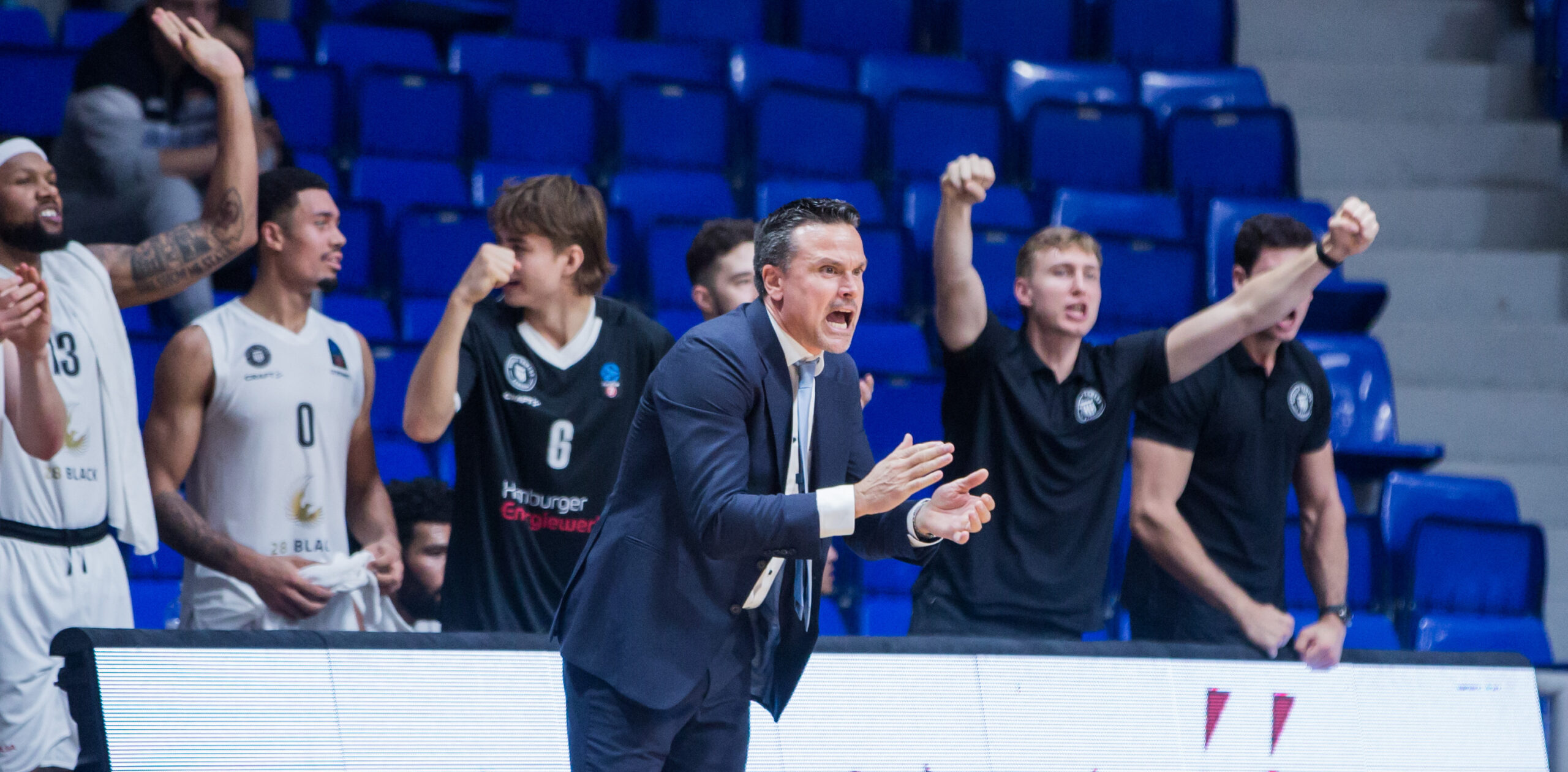 Basketballtrainer in Anzug gestikuliert energisch, Spieler und Teammitglieder jubeln im Hintergrund auf der Bank