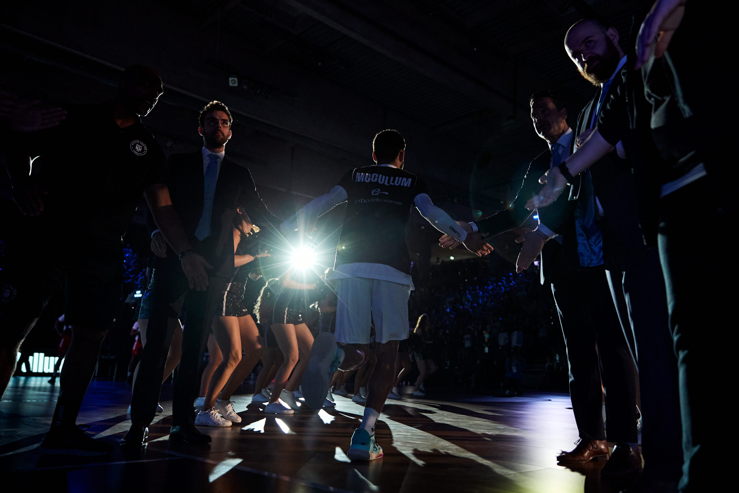 Basketballspieler mit dem Namen 'McCollum' auf dem Trikot läuft durch eine Reihe von Menschen, die Hände zum Abklatschen ausstrecken, beleuchtet von starkem Licht im Hintergrund