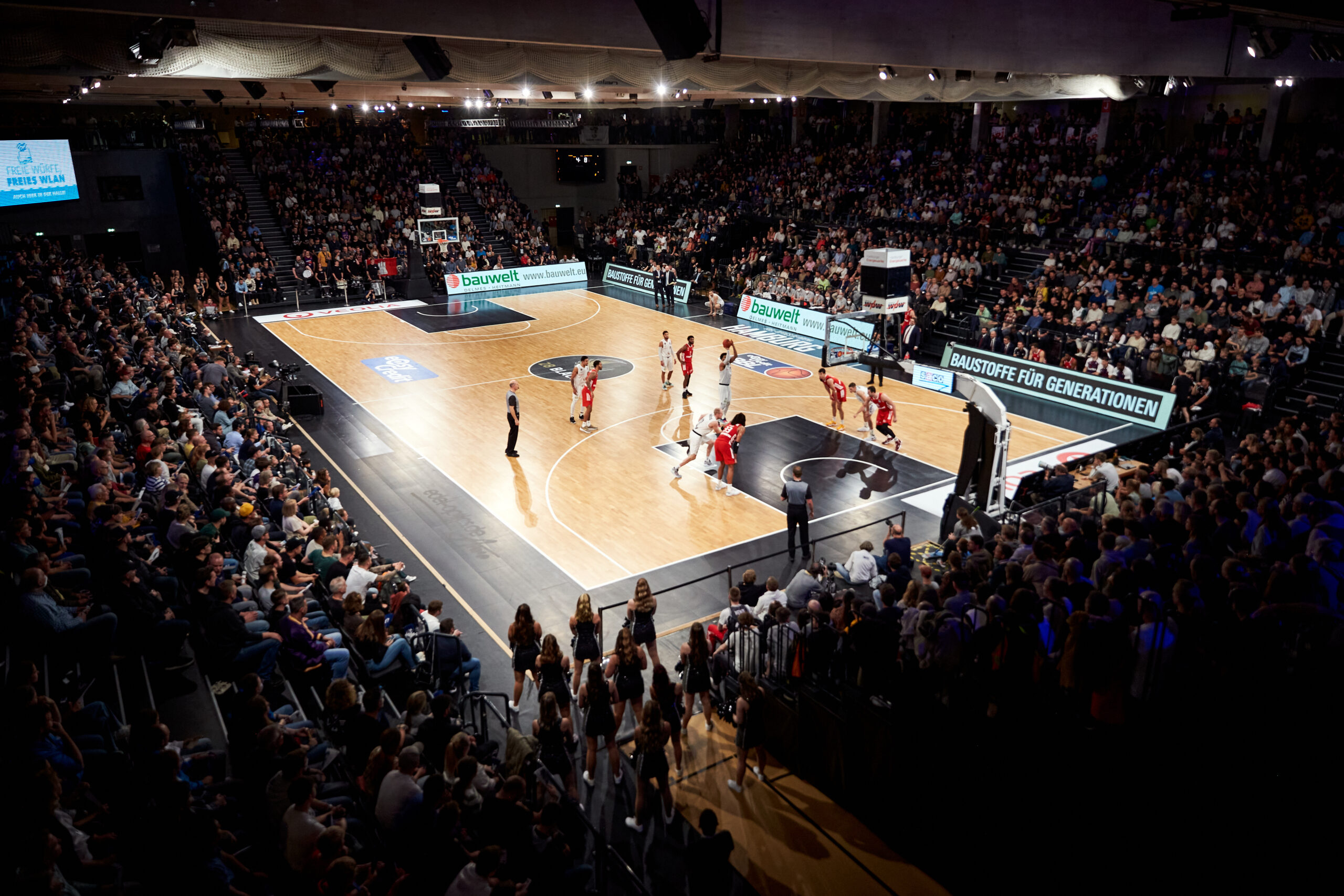 Basketballspiel in einer vollen Halle mit Spielern in weißen und roten Trikots, Schiedsrichter und jubelndem Publikum