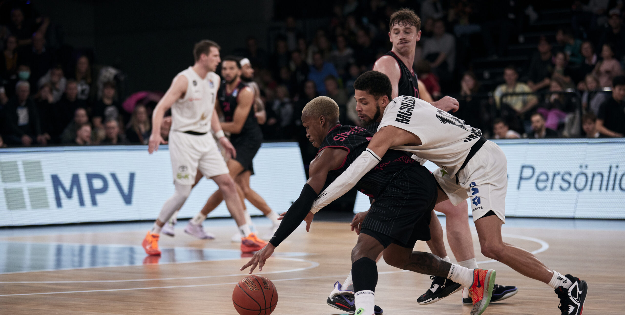 Zwei Basketballspieler kämpfen um den Ball auf dem Spielfeld, umgeben von weiteren Spielern und Zuschauern im Hintergrund.