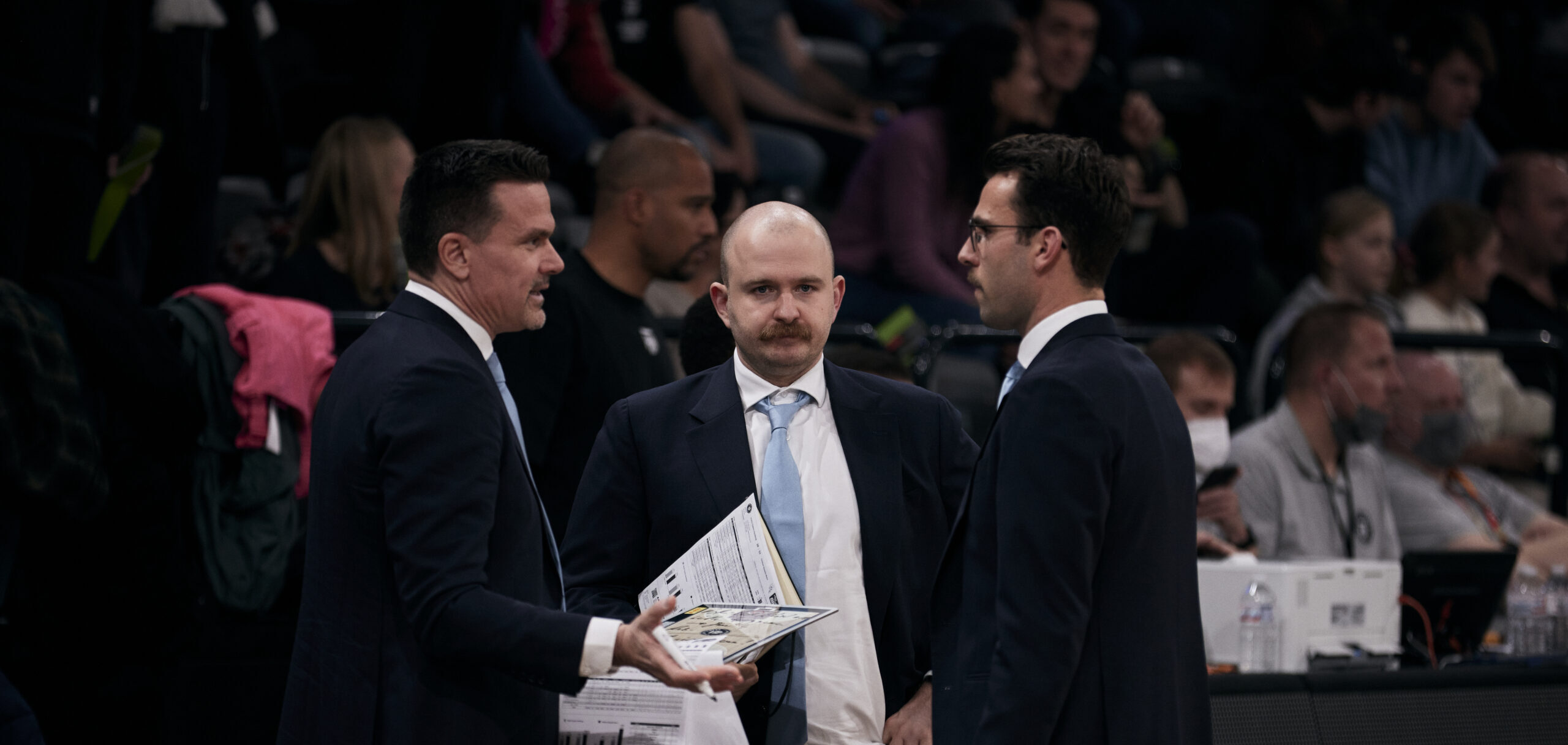 Drei Männer in Anzügen diskutieren während eines Basketballspiels, einer hält Spielpläne in der Hand