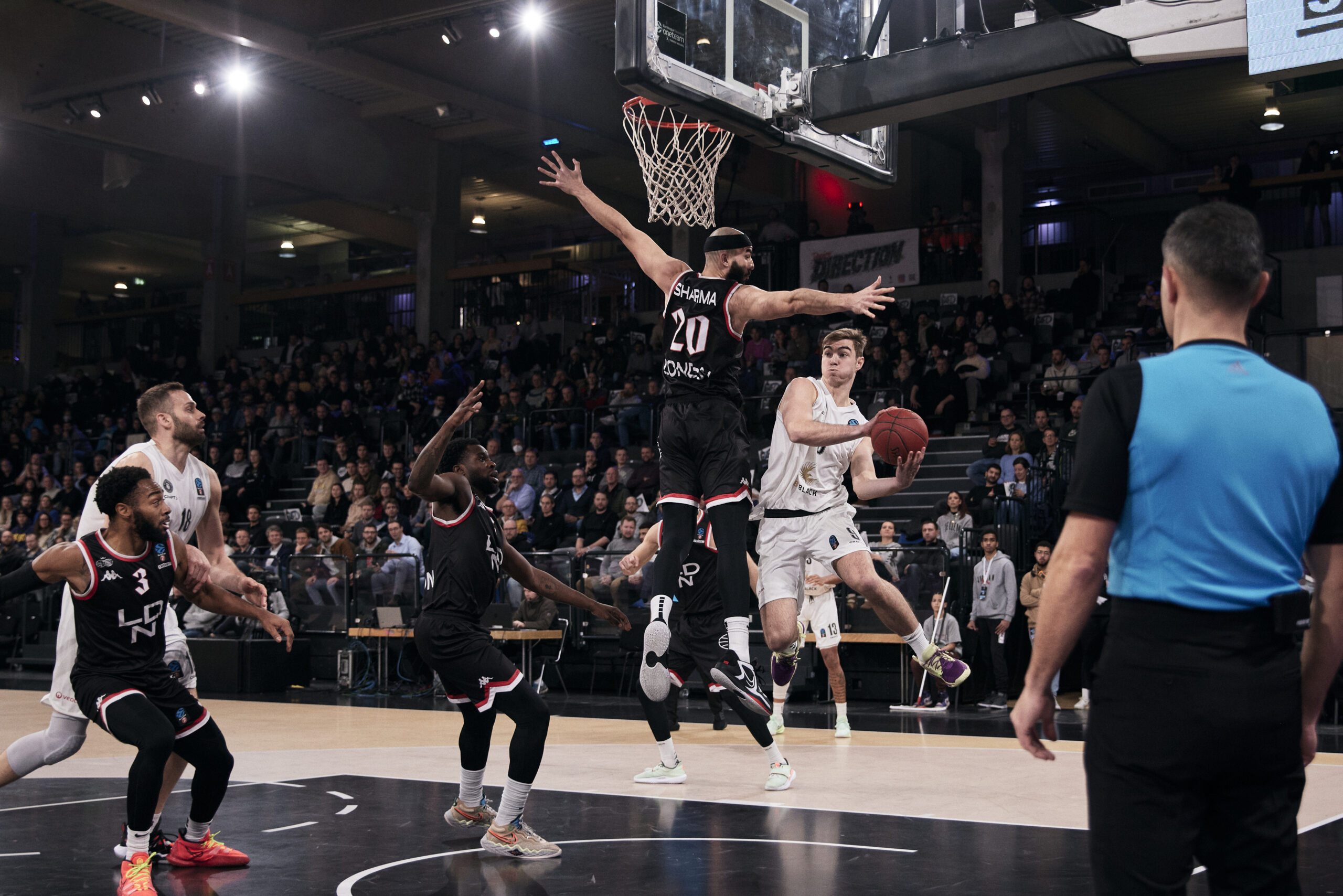 Basketballspieler in weißer Uniform springt zum Korb, von mehreren Spielern in schwarzen Trikots verteidigt, Schiedsrichter im Vordergrund, Publikum im Hintergrund