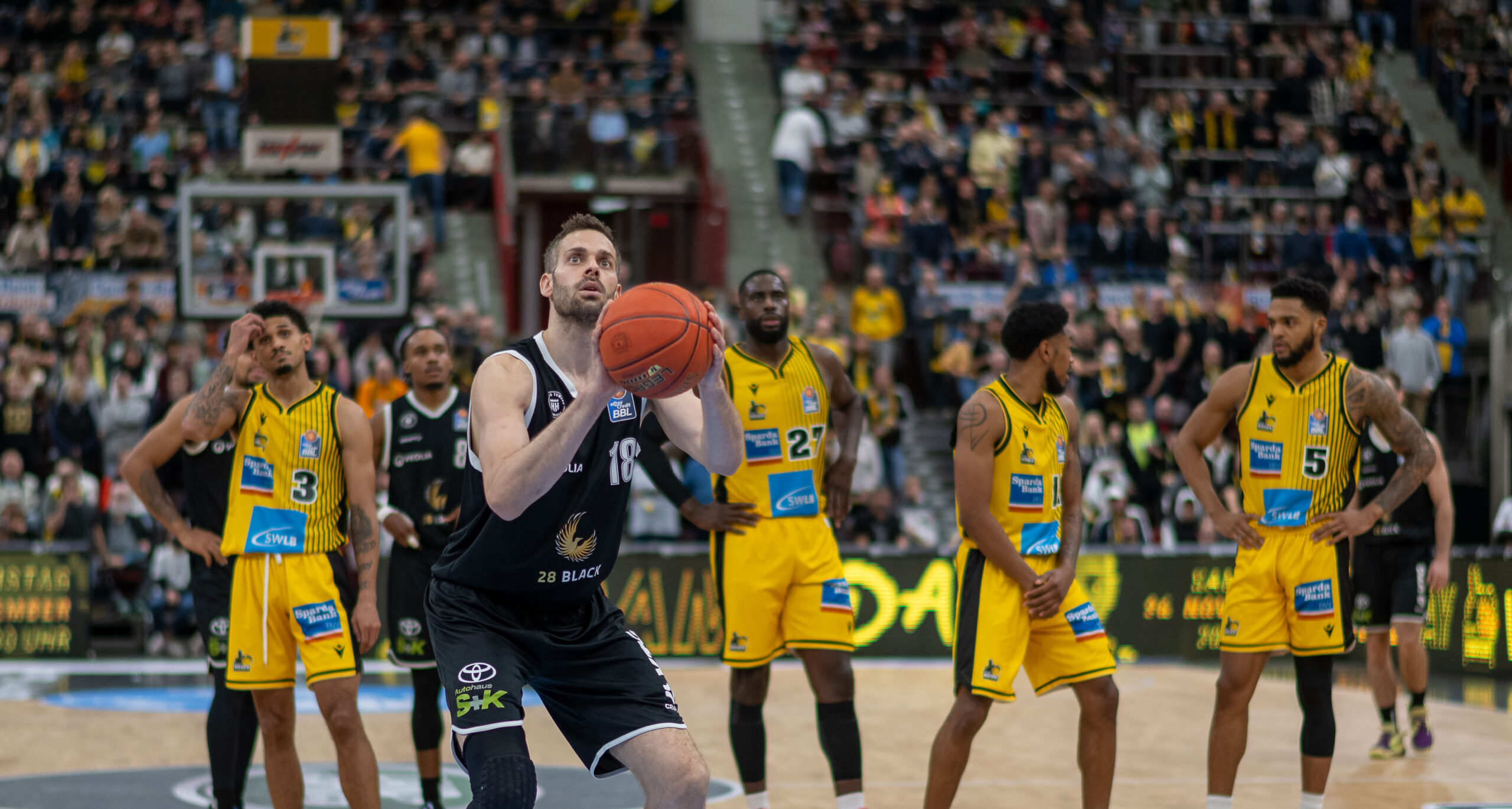Basketballspieler in schwarzem Trikot macht Freiwurf, umgeben von Spielern in gelben Trikots auf dem Spielfeld