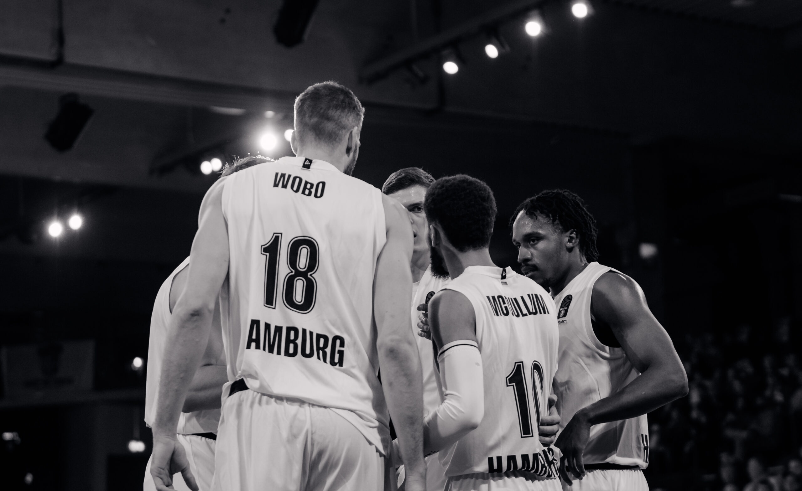 Basketballspieler in weißen Trikots mit den Namen 'Wobo' und 'McCullum' in einer Spielpause, sie diskutieren im Teamkreis
