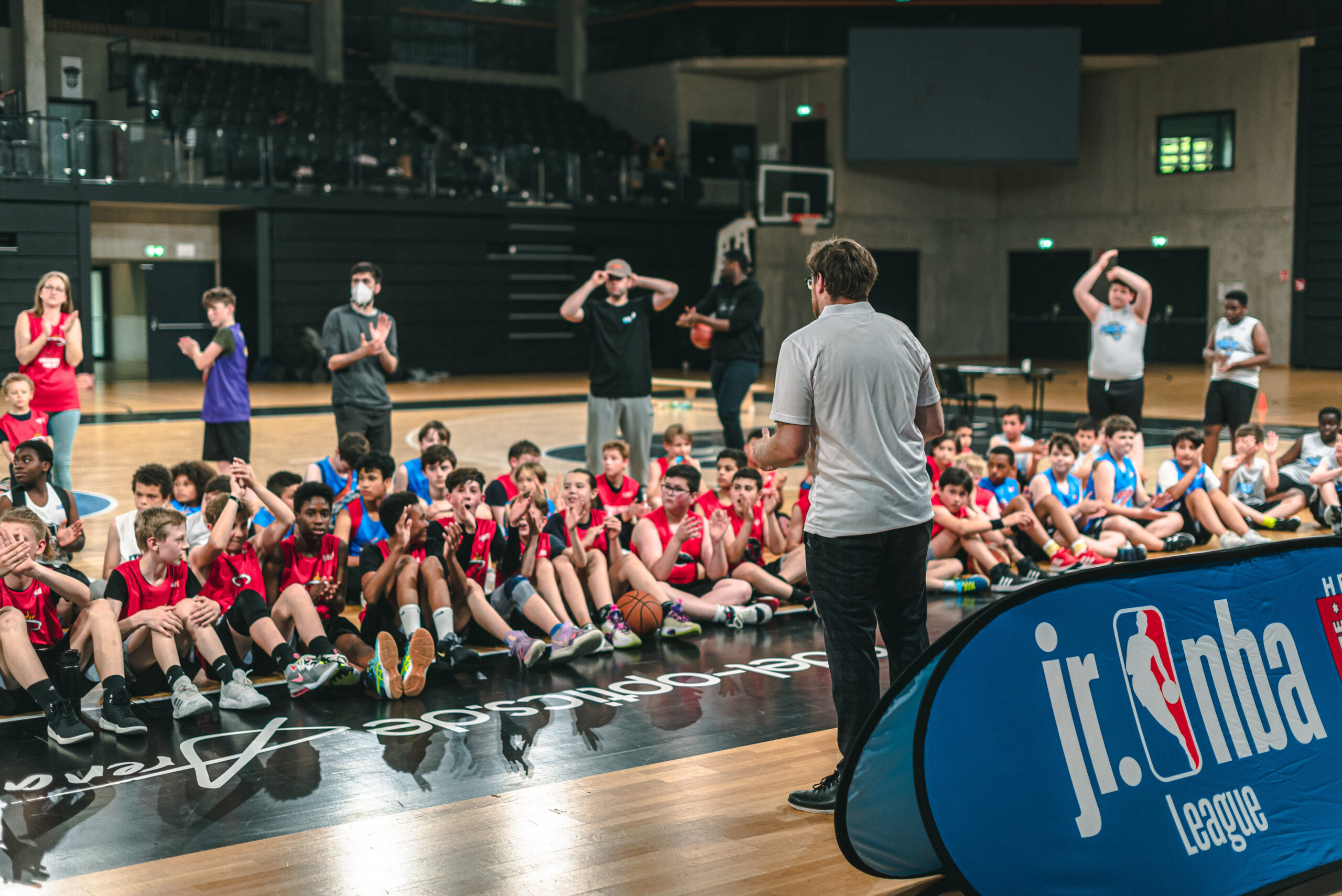 Trainer spricht zu einer Gruppe von Kindern in Basketballkleidung, die auf dem Hallenboden sitzen, neben einem Banner mit dem Schriftzug 'jr.nba League'
