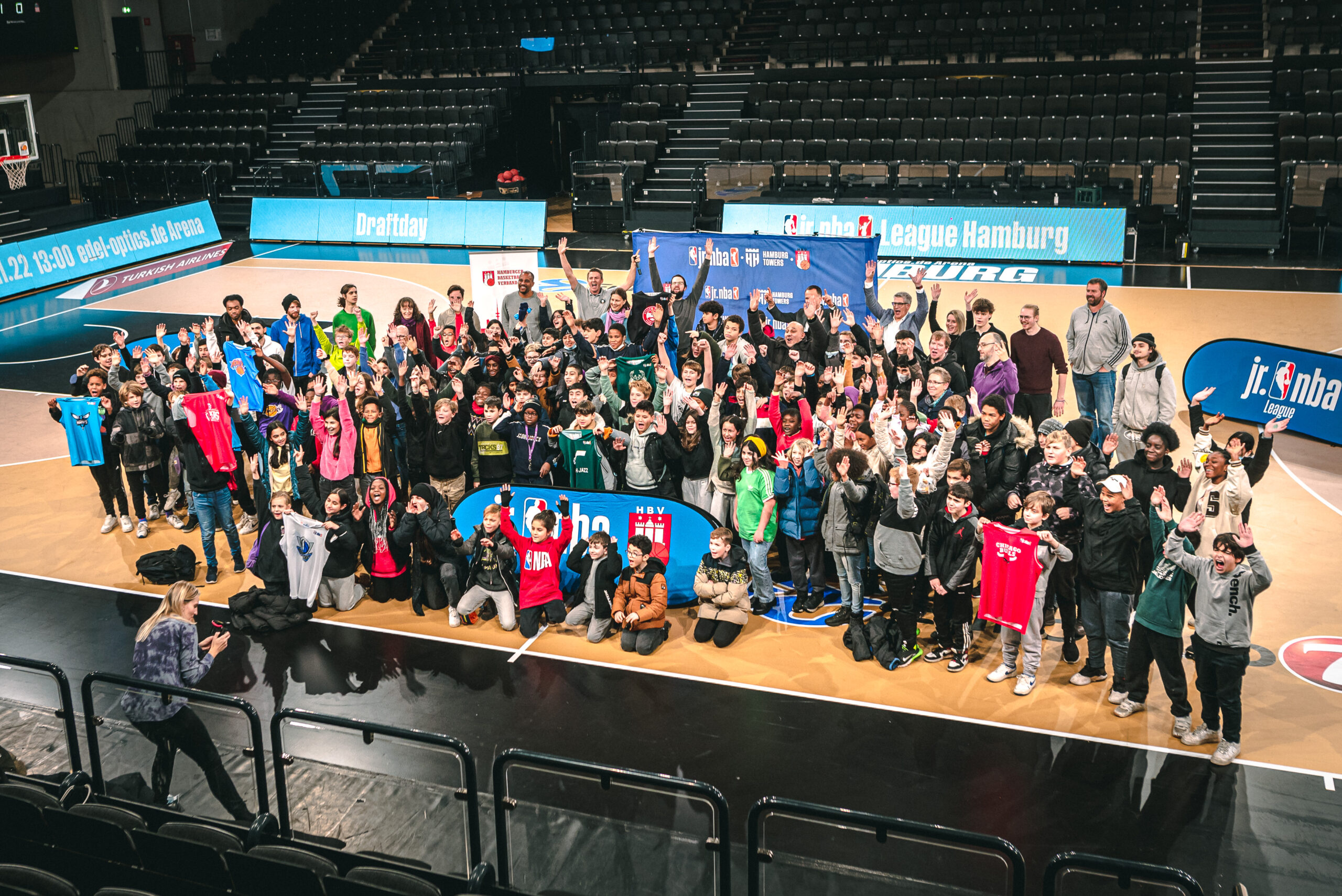 Große Gruppe von Kindern und Jugendlichen auf einem Basketballfeld, viele halten jr. NBA-Trikots, im Hintergrund Banner mit 'jr. NBA League Hamburg' und 'Draftday'