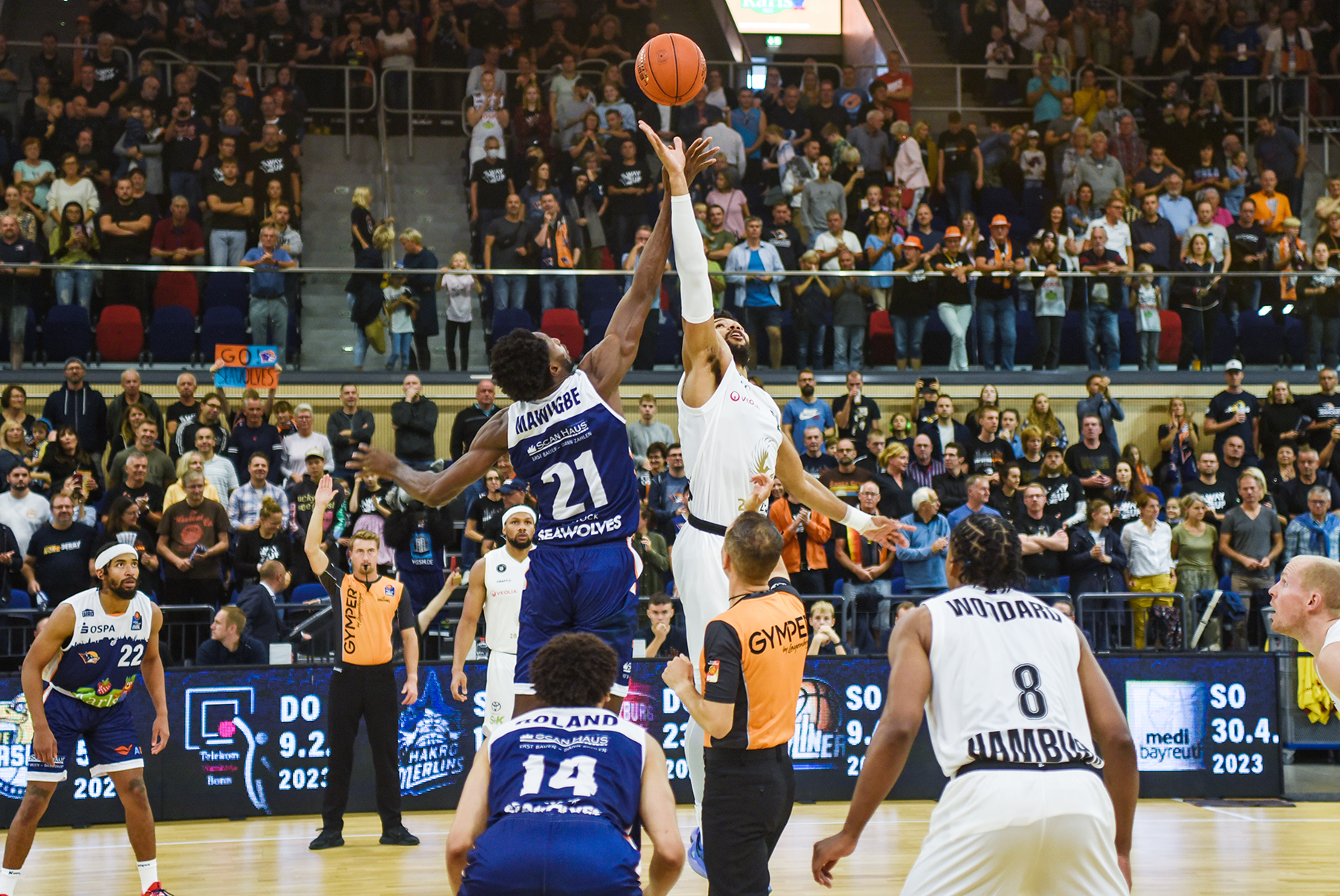 Zwei Basketballspieler beim Sprungball, umgeben von Spielern und Publikum in der Halle