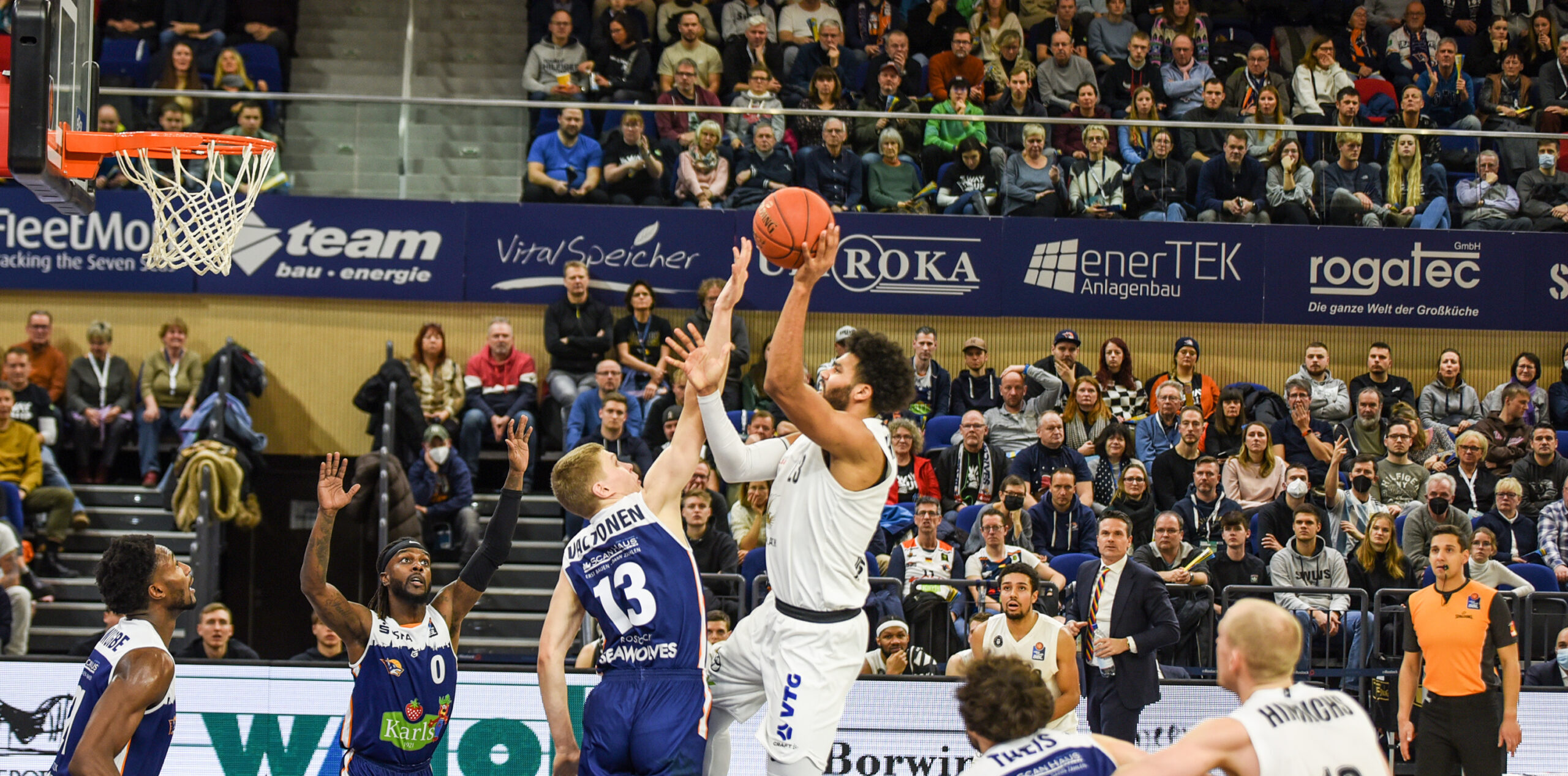 Basketballspieler in weißem Trikot springt zum Korb, verteidigt von Spieler in blauem Trikot mit Nummer 13, Zuschauer im Hintergrund