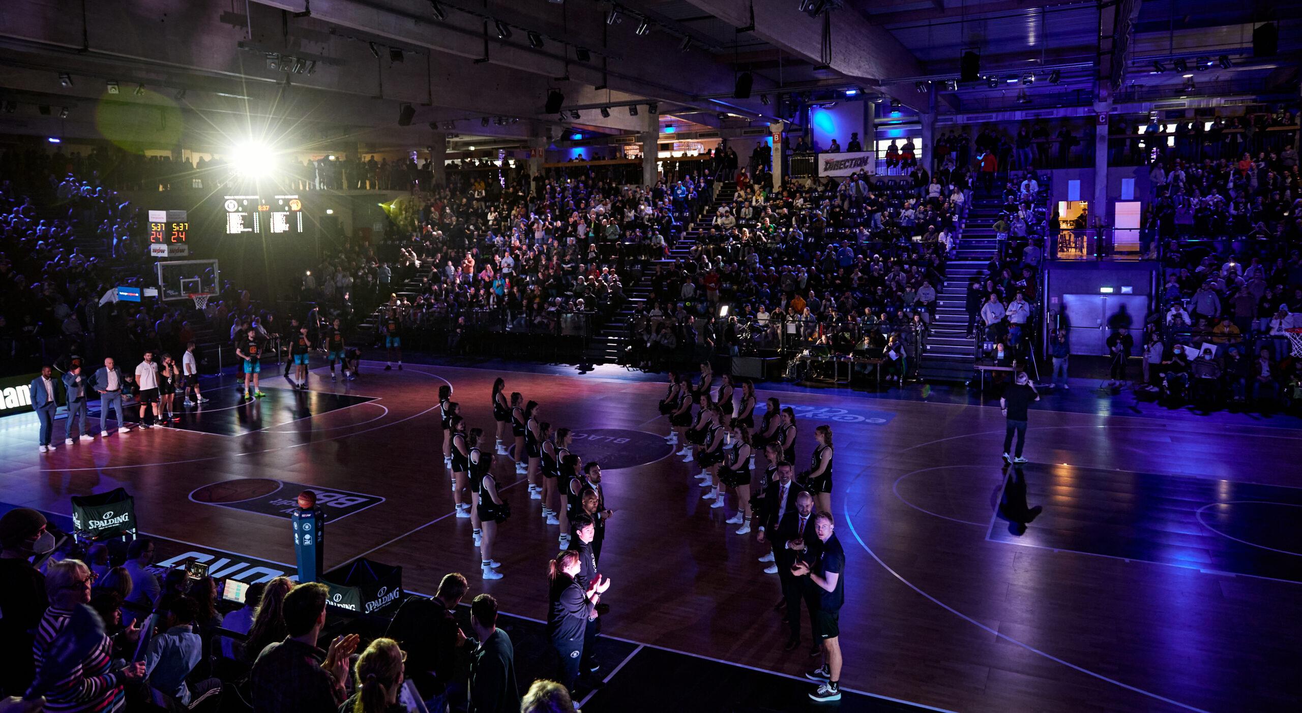 Basketballspiel in einer Halle mit Zuschauern auf den Tribünen, Spieler und Cheerleader auf dem Spielfeld bei der Vorstellung