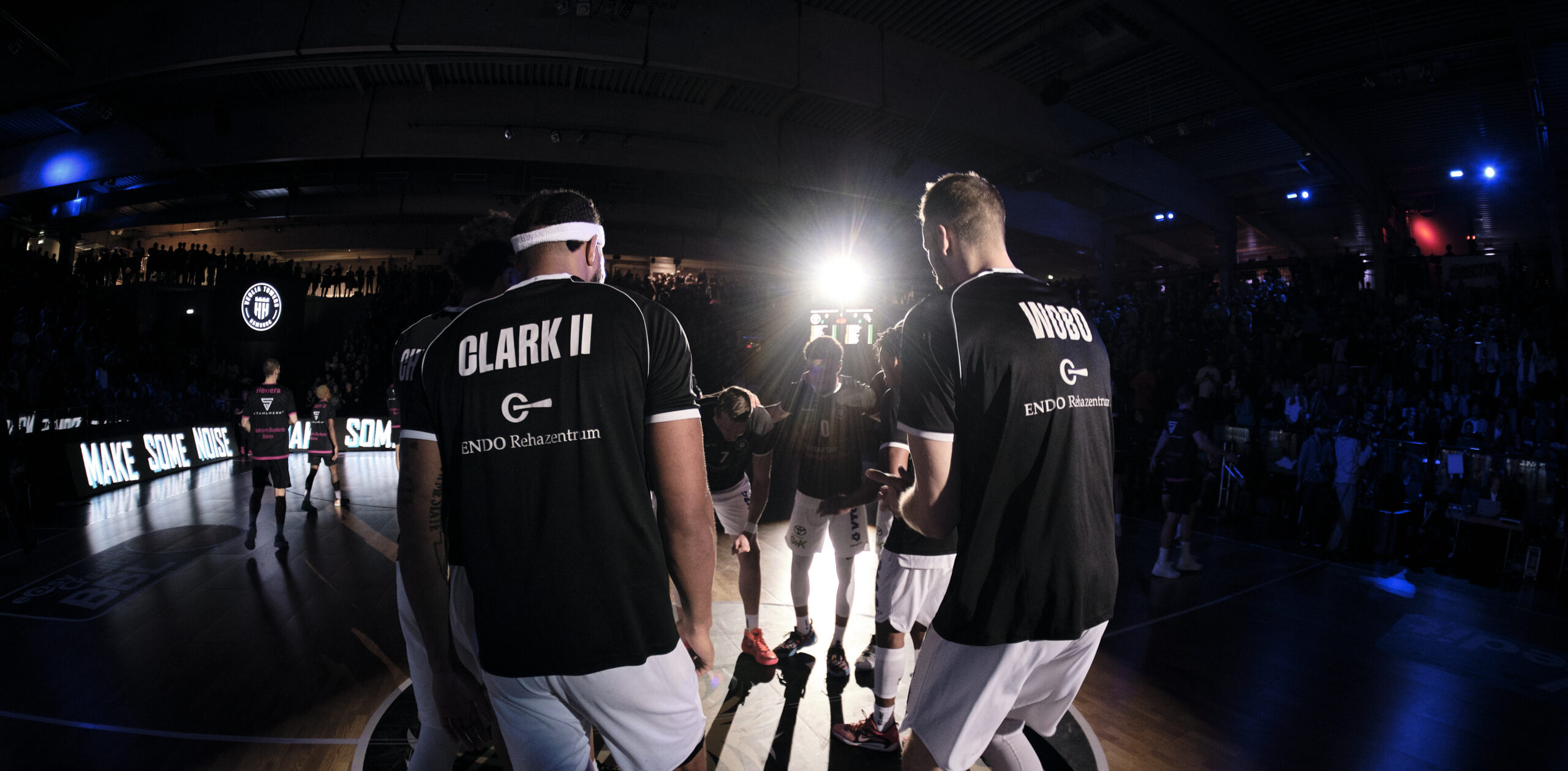 Basketballspieler Clark II und Wood vom Team Towers stehen vor dem Spiel in der Halle, Zuschauer im Hintergrund