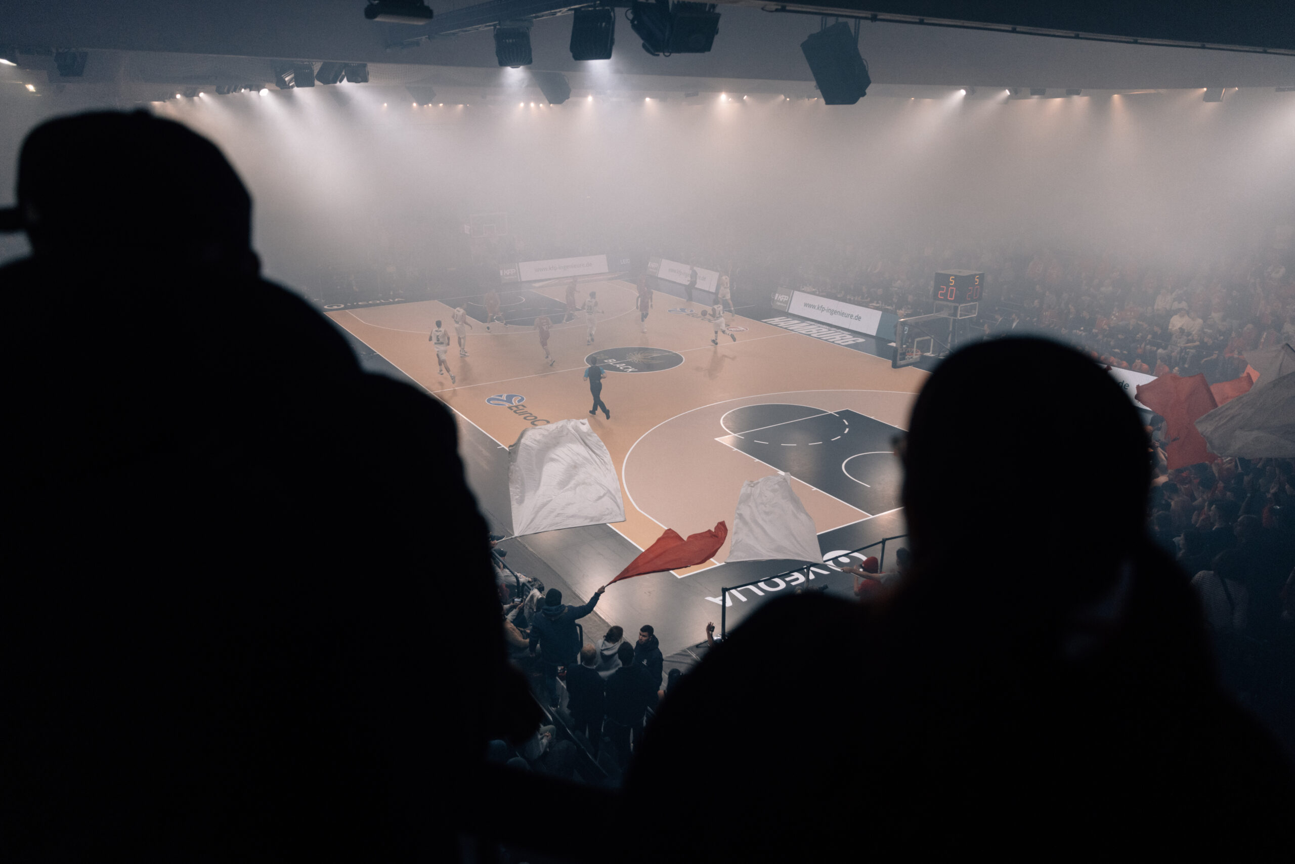 Blick von den Rängen auf ein Basketballspiel in einer Halle mit Spielern auf dem Spielfeld und Zuschauern im Hintergrund