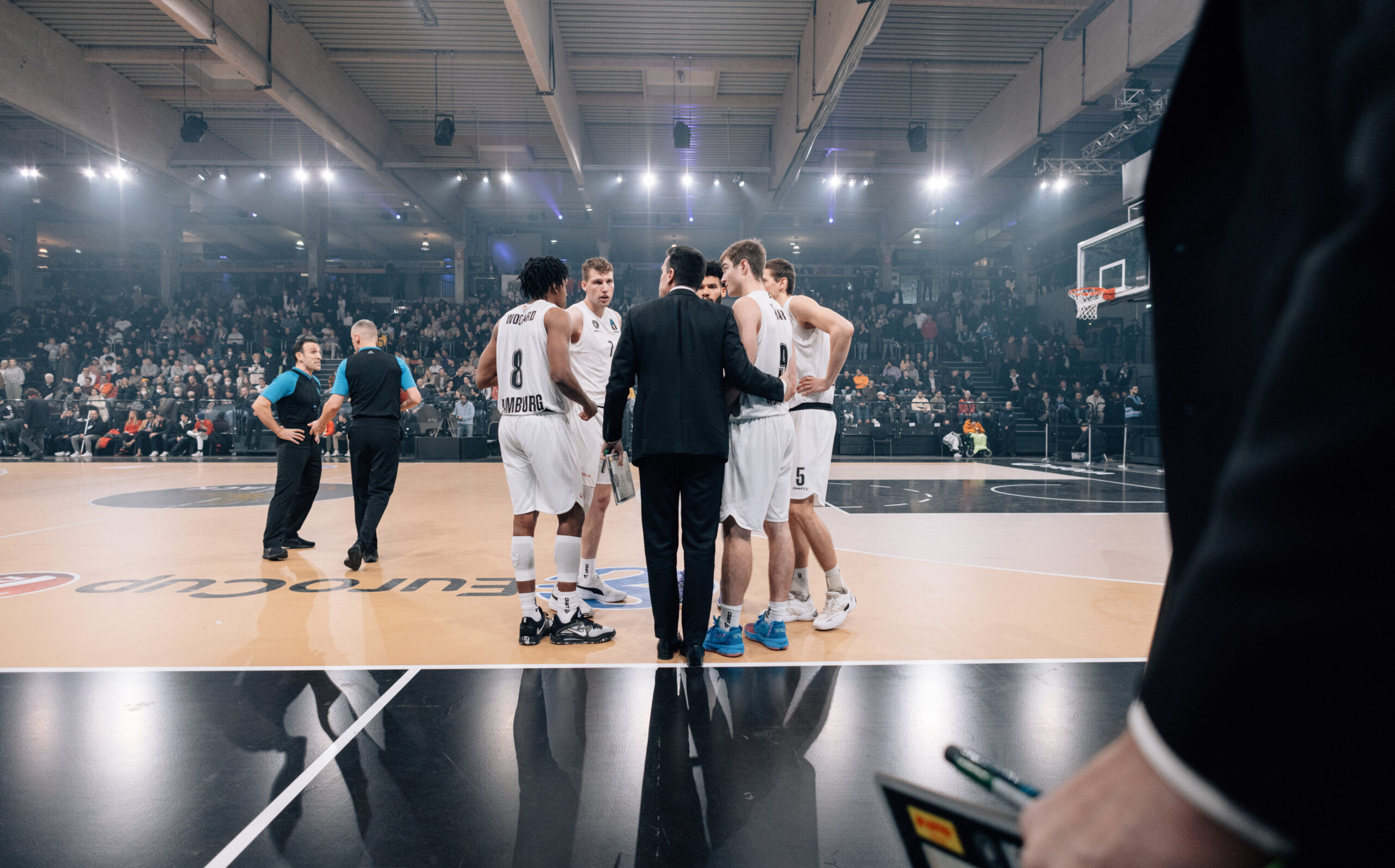 Basketballspieler in weißen Trikots und Trainer in der Spielfeldmitte während einer Auszeit in einer vollen Halle