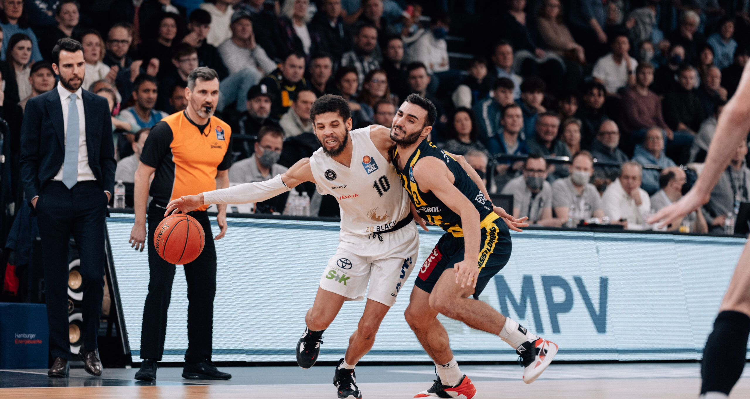 Zwei Basketballspieler kämpfen um den Ball, einer in weißem Trikot mit der Nummer 10, der andere in dunklem Trikot mit gelben Akzenten, Zuschauer im Hintergrund
