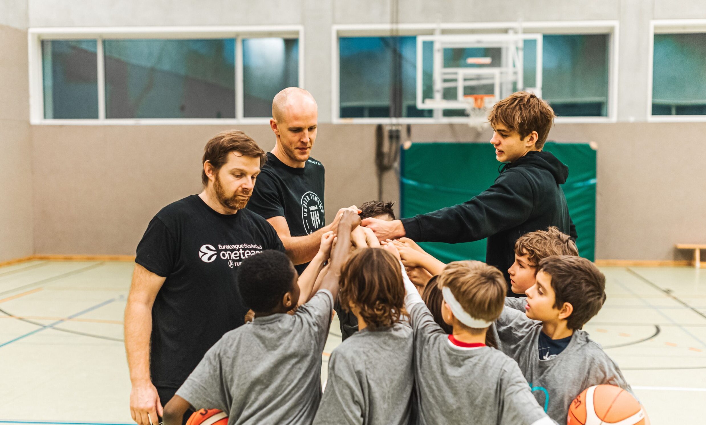Jugendliche Basketballspieler und Trainer im Kreis, Hände zusammengelegt in einer Sporthalle
