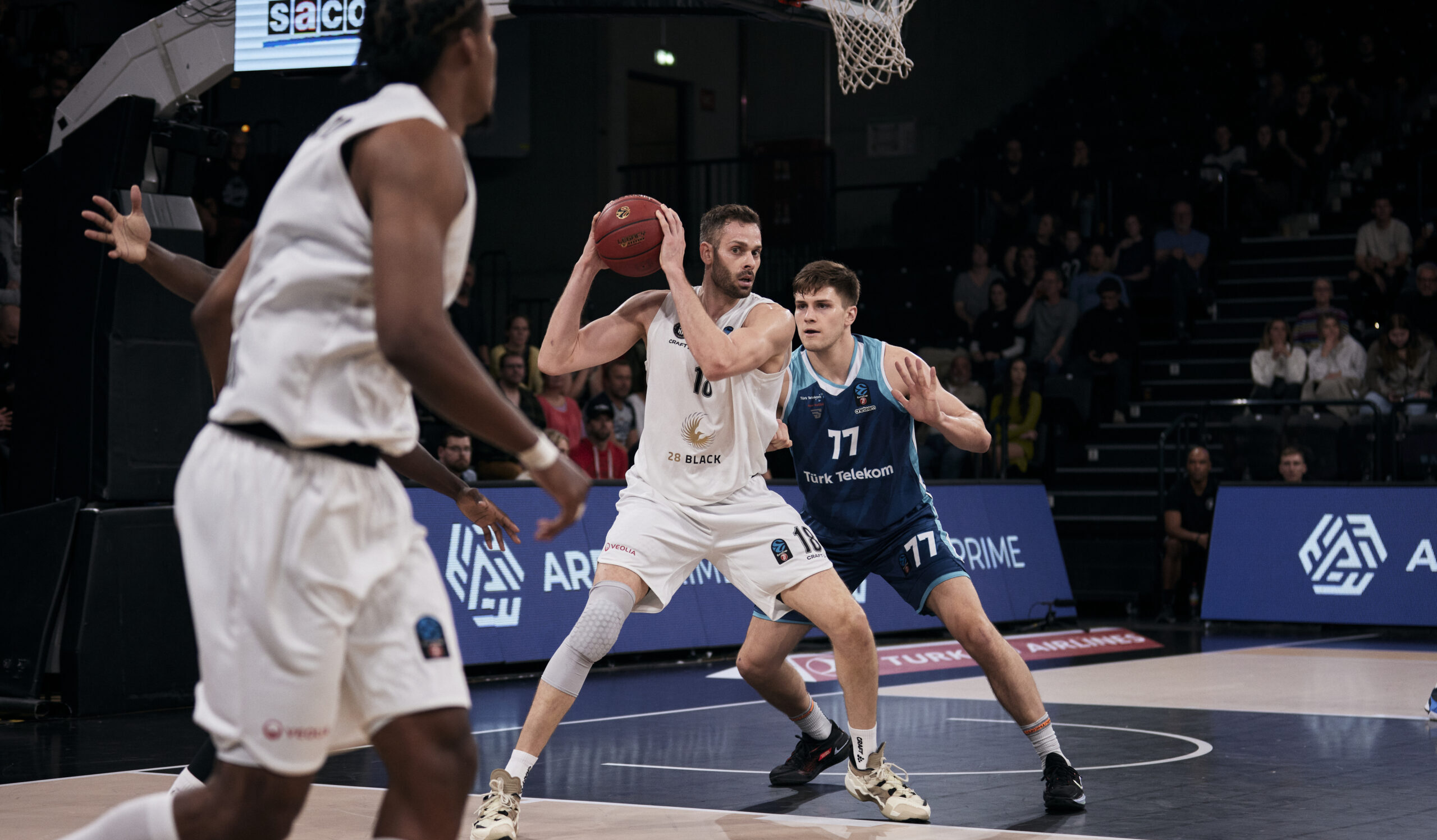 Basketballspieler in weißer Uniform hält Ball, wird von zwei Spielern in blauen Trikots verteidigt, Spiel in Halle mit Zuschauern