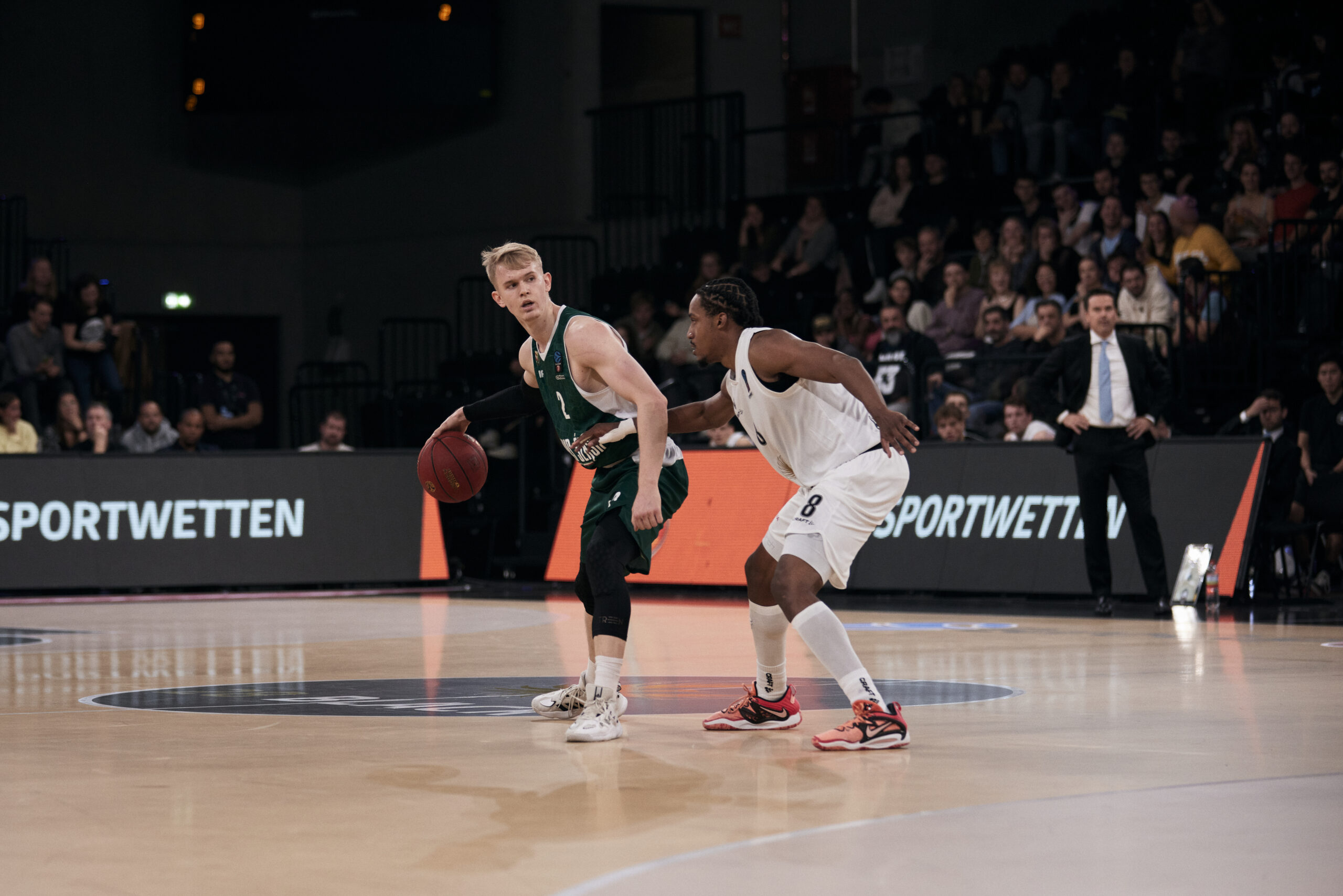 Zwei Basketballspieler auf dem Spielfeld, einer in grün mit Ball, der andere in weiß verteidigt, Zuschauer im Hintergrund