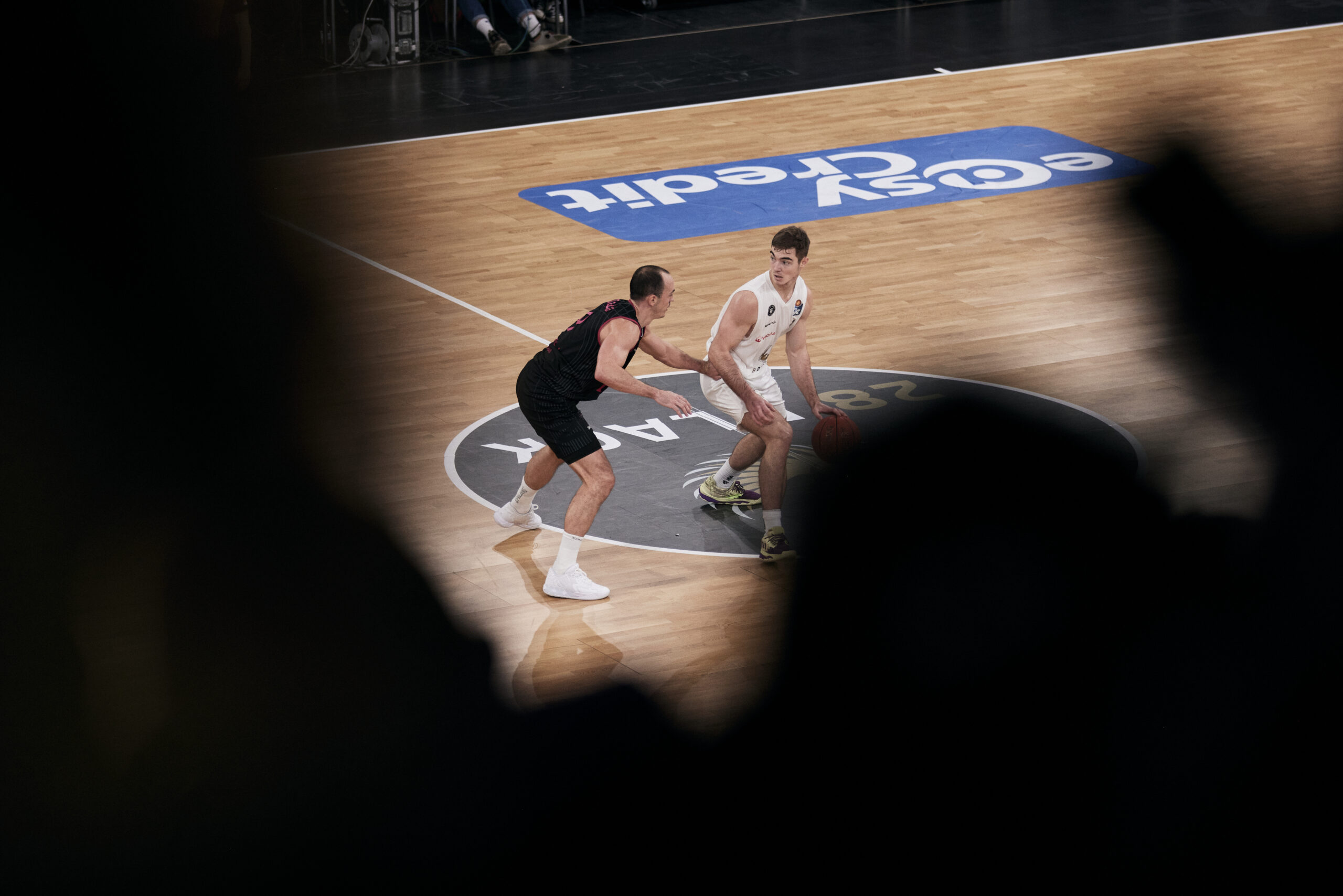 Zwei Basketballspieler auf dem Spielfeld, einer in weiß mit Ball, der andere in schwarz verteidigt ihn
