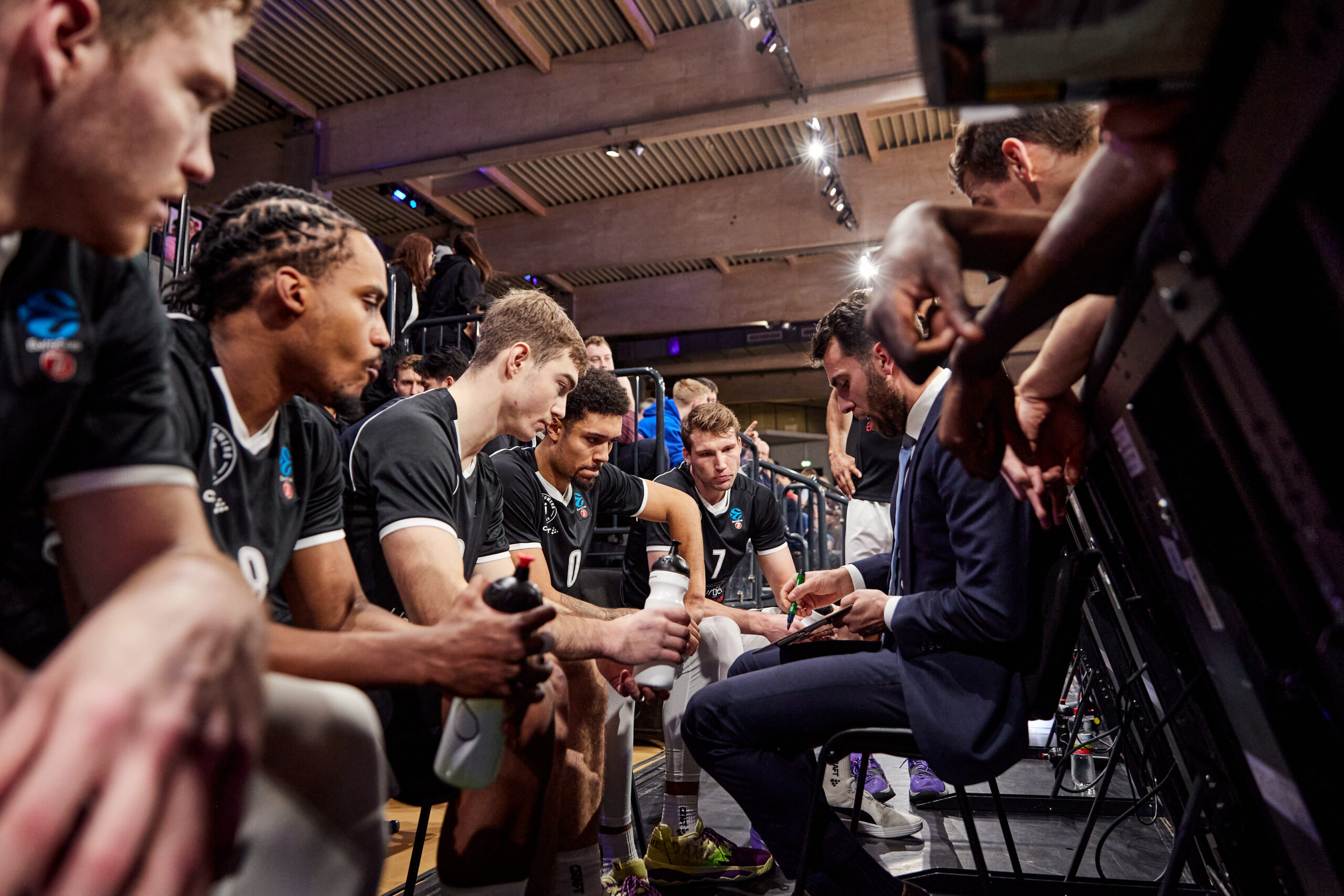 Basketballspieler der Towers in schwarzen Trikots sitzen auf einer Bank und hören einem Trainer zu, der Anweisungen gibt