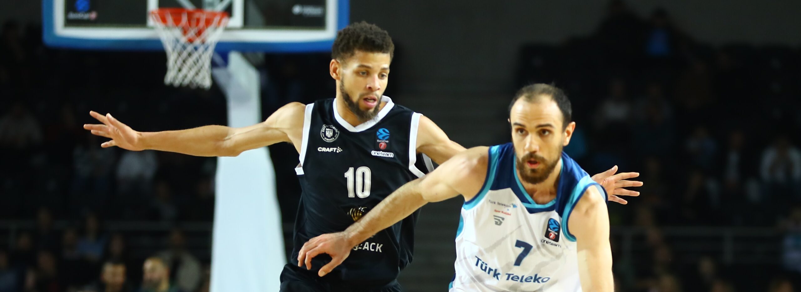 Zwei Basketballspieler in Aktion, einer in schwarzem Trikot mit Nummer 10, der andere in weiß-blauem Trikot, kämpfen um den Ball vor dem Korb.