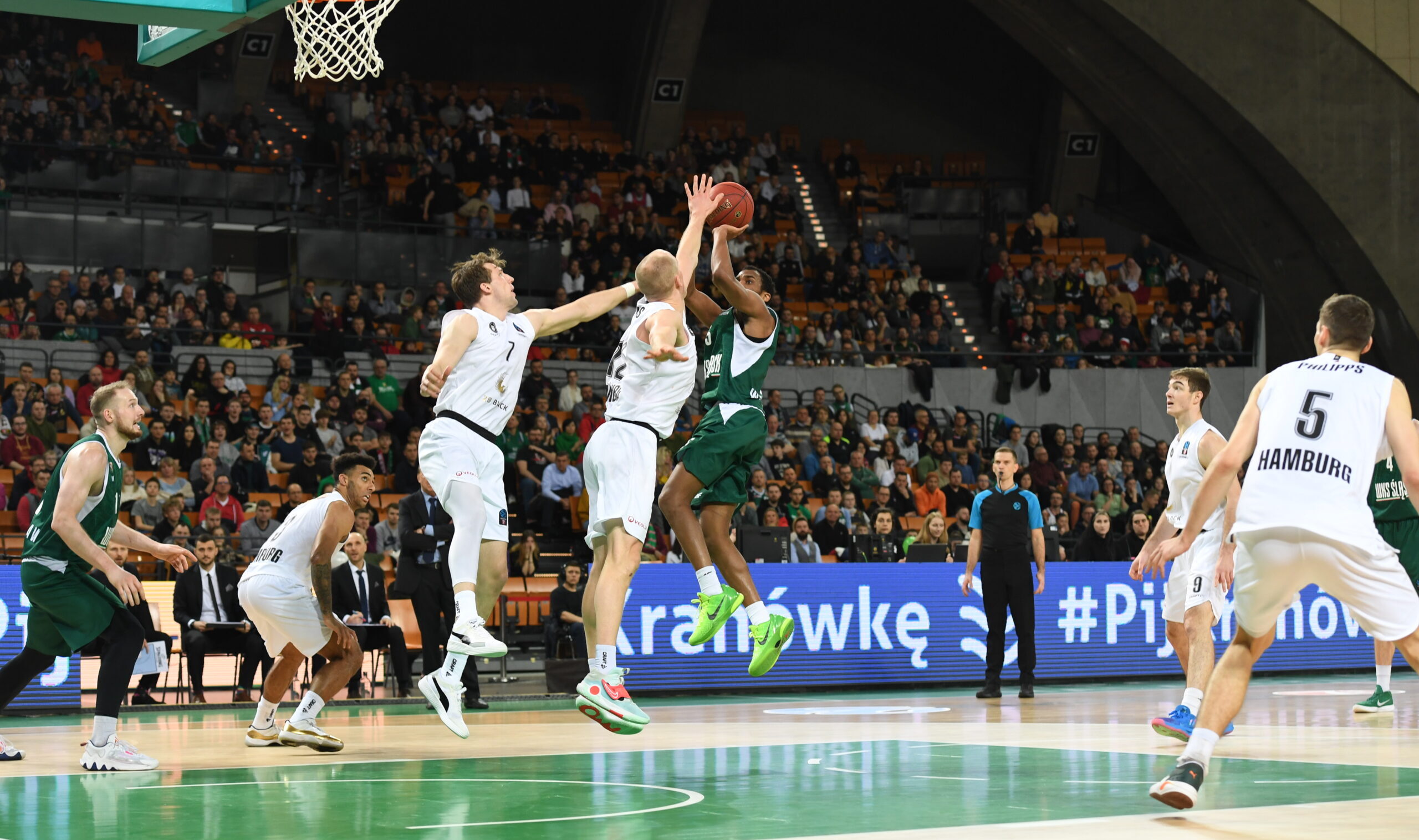 Basketballspiel zwischen zwei Teams, ein Spieler in grün springt zum Korb, mehrere Spieler in weiß verteidigen aktiv