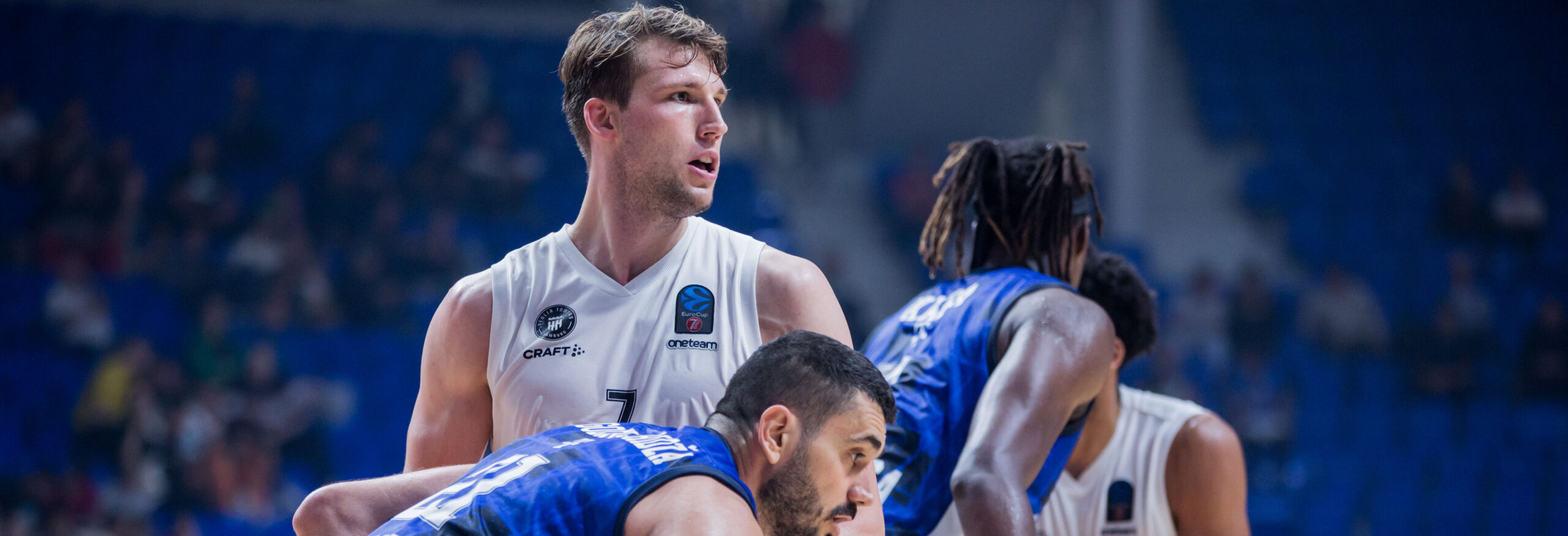 Zwei Basketballspieler in weißen Trikots und zwei in blauen Trikots kämpfen um den Ball während eines Spiels in einer Halle