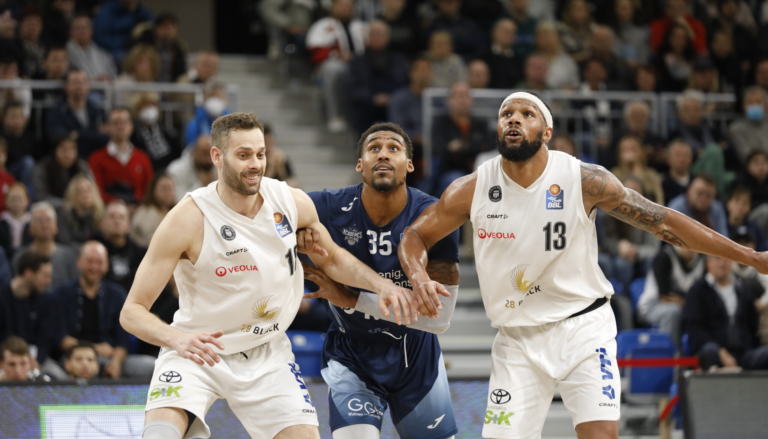Drei Basketballspieler in Aktion, zwei in weißen Trikots, einer in blauem Trikot mit der Nummer 35, auf einem Spielfeld vor Zuschauern