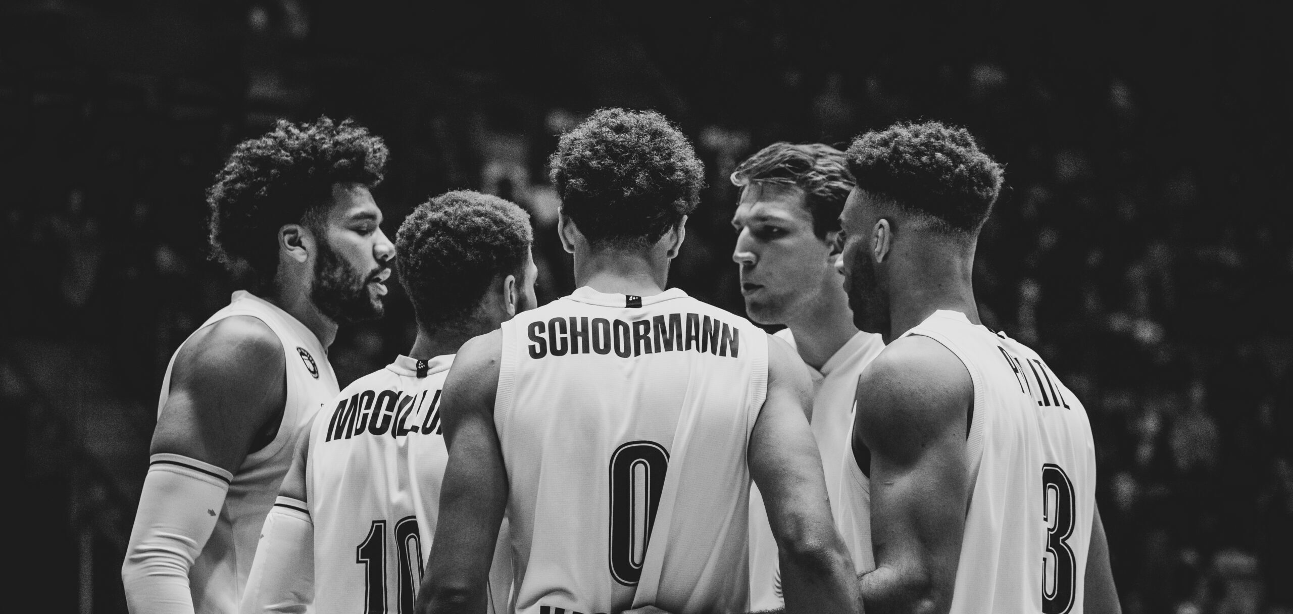 Fünf Basketballspieler in einem Teamkreis, tragen Trikots mit den Namen Schoormann, McClung und Jeter, konzentrieren sich auf das Spiel.