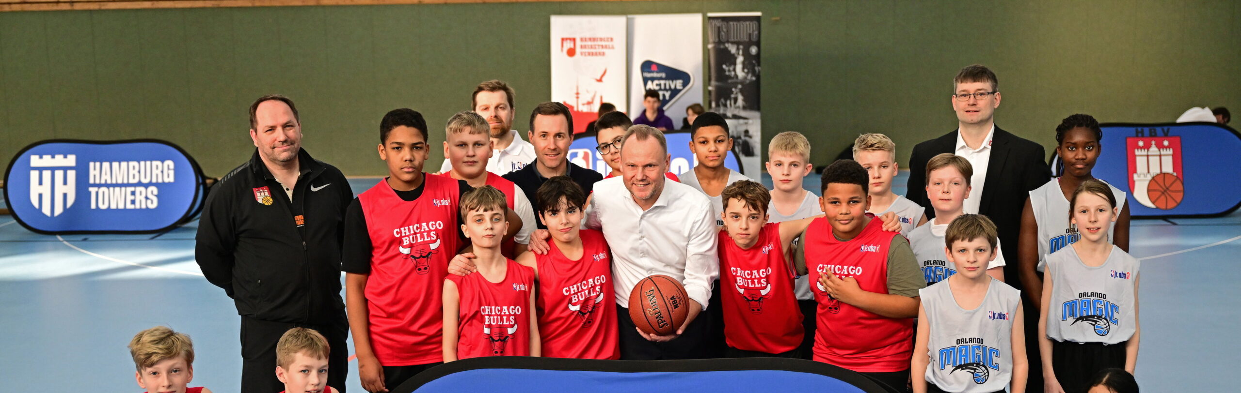 Jugendliche Basketballspieler in roten und weißen Trikots posieren in einer Sporthalle mit zwei Trainern